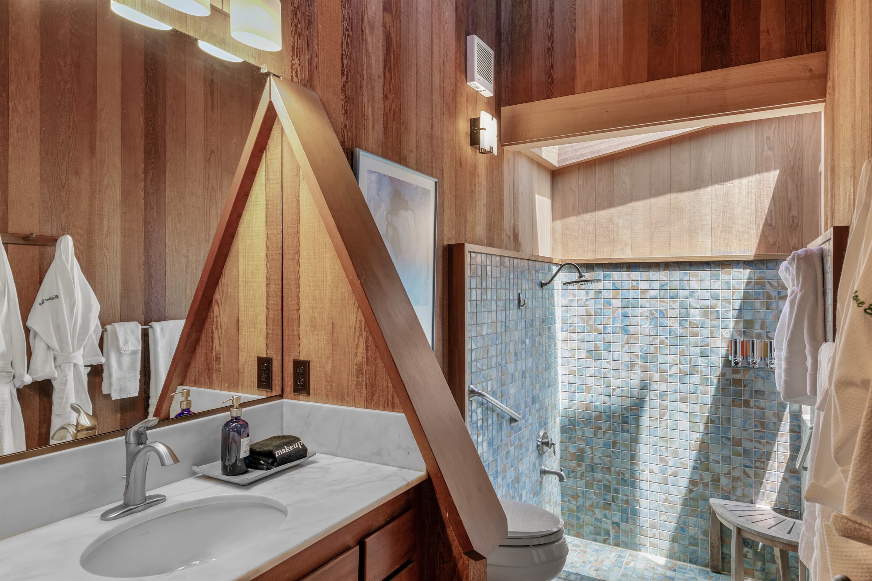 Elegant ensuite bath—offering a serene, spa-like experience with refined coastal charm.  Features wood-paneled walls, shower head, plush robes, and a walk-in shower with mosaic tile and natural light