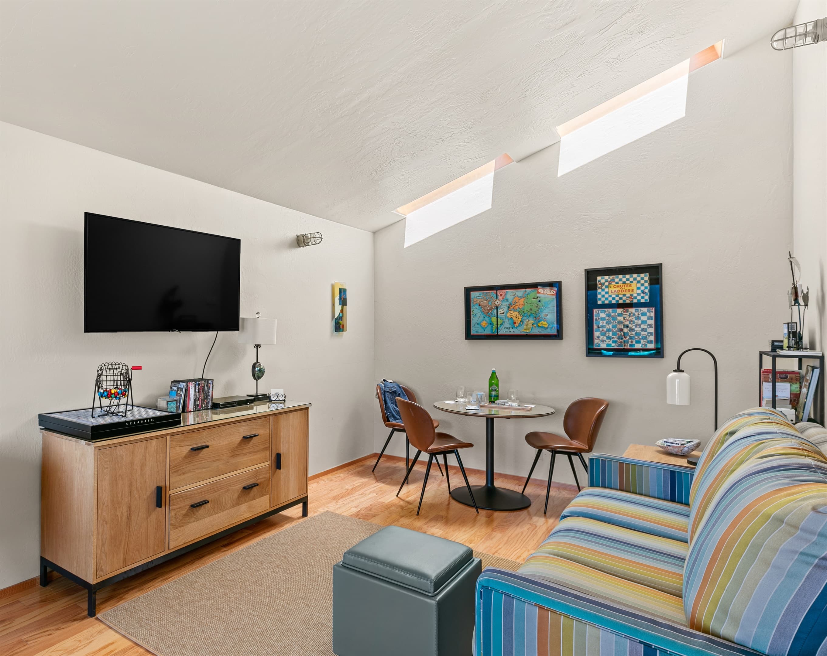 Bright, stylish game room featuring skylights with blinds, a colorful striped sleeper sofa, flat-screen TV, and round dining table—an inviting, upscale space for games, movies, and leisurely gatherings. 