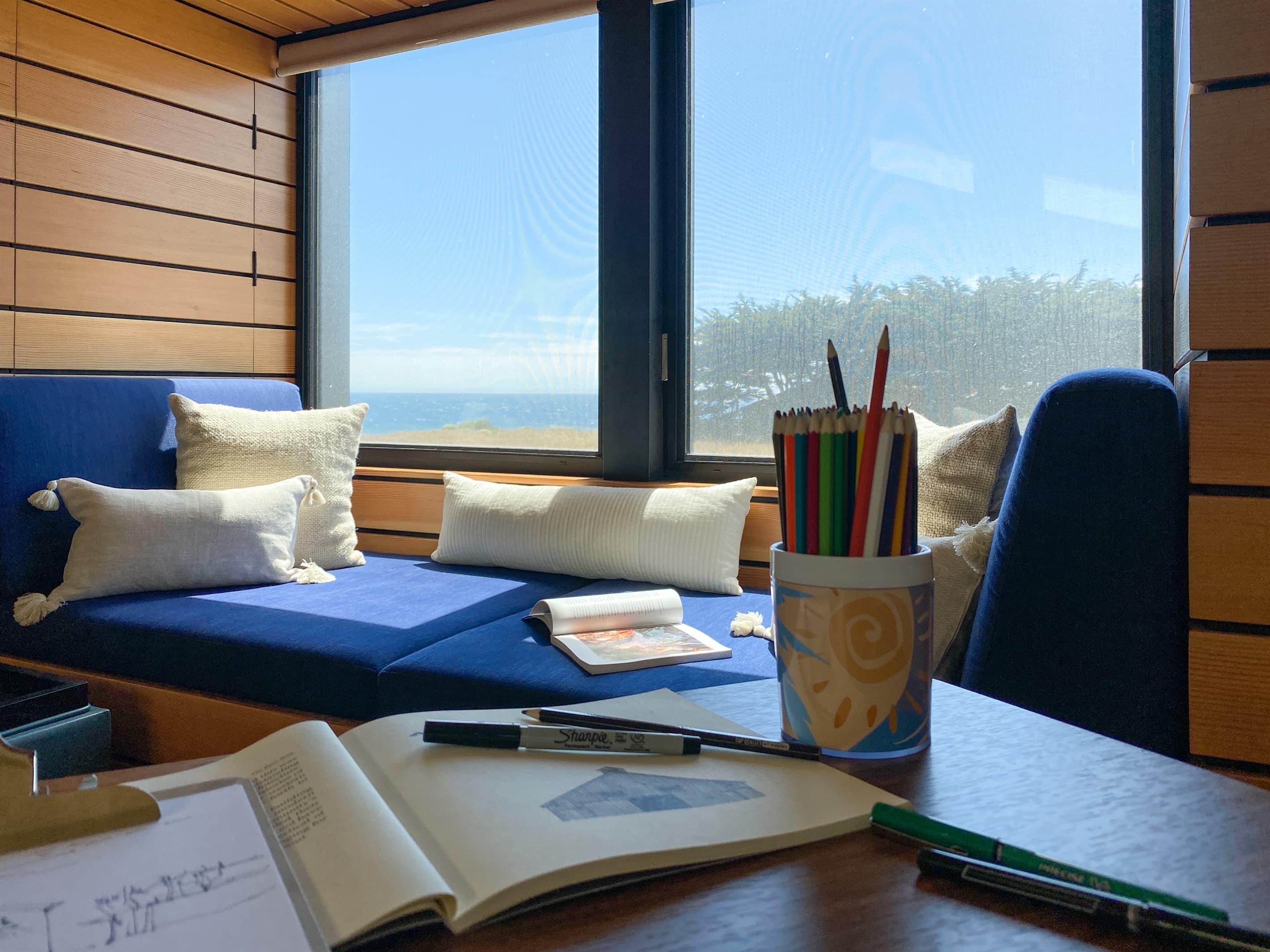 Coastal creativity meets comfort: A sunlit window seat with ocean views, cozy pillows, and art supplies invites peaceful sketching, reading, or daydreaming—an inspiring nook for refined, restful moments. Sketch/journaling booklets provided.
