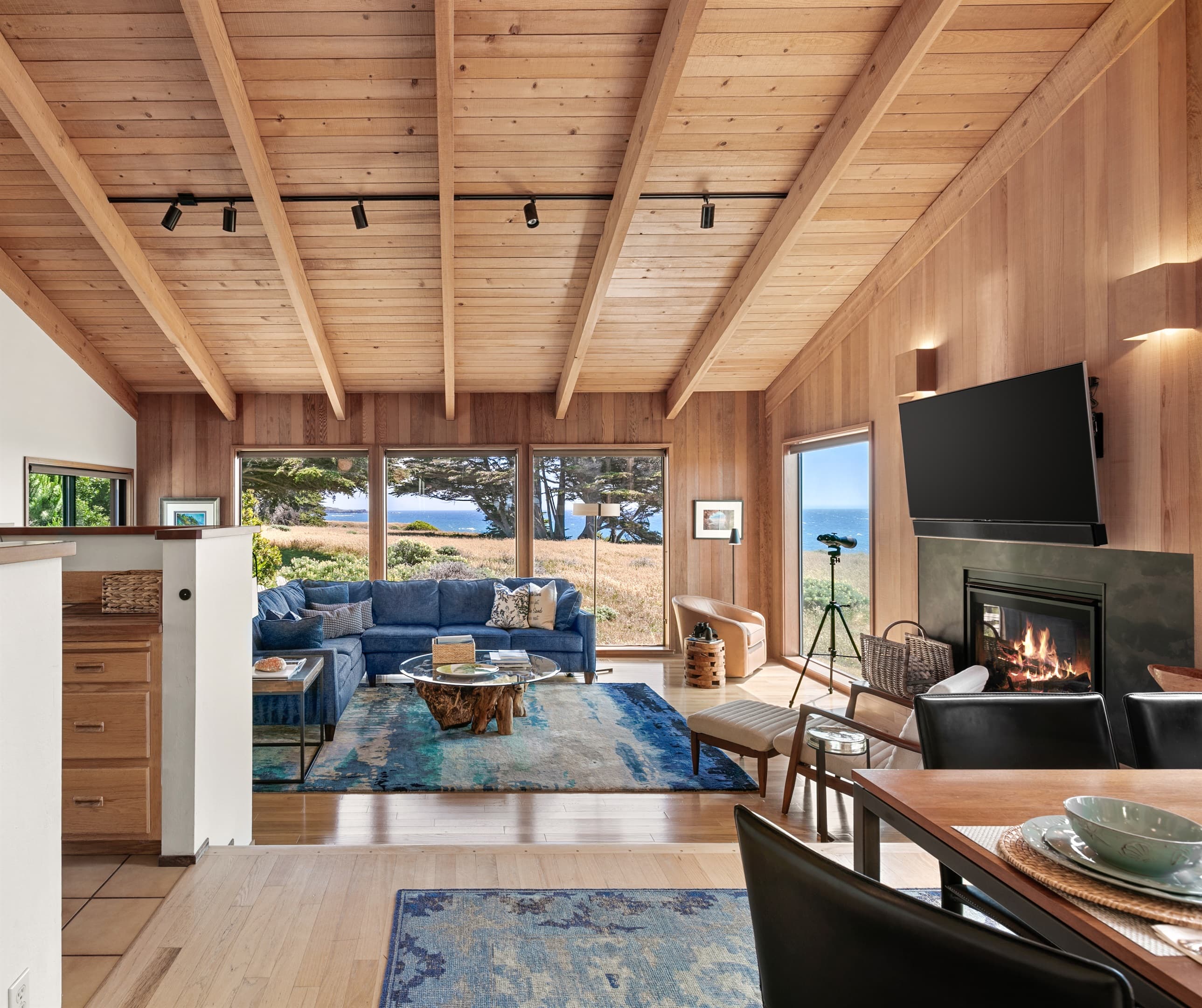 Sophisticated coastal living room with vaulted wood ceilings, fireplace, and ocean-view windows. Layered blue-toned area rugs echo the sea, adding warmth and texture to this refined retreat inspired by nature.