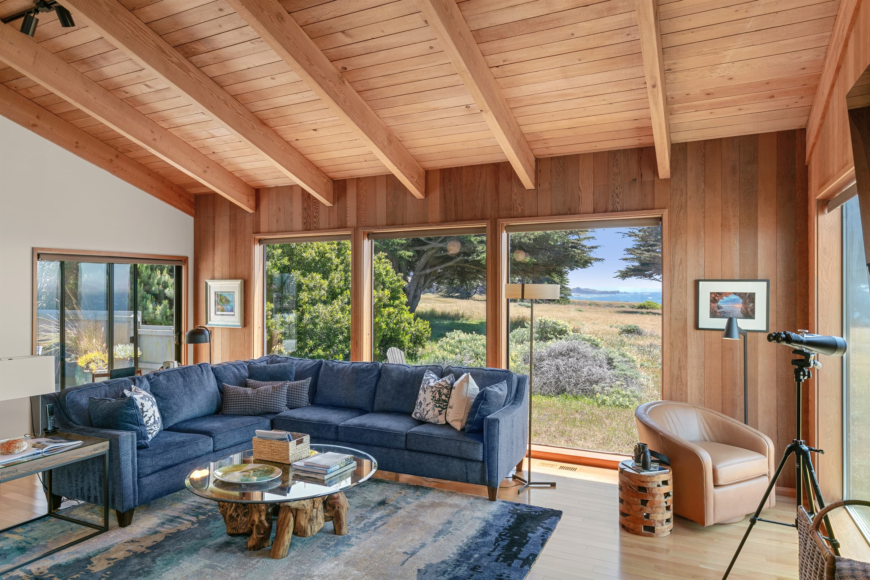 Sun-drenched living room with vaulted wood ceilings, expansive windows framing ocean and meadow views, plush sectional, and nature-inspired textures—offering refined comfort and coastal elegance in every detail. 
