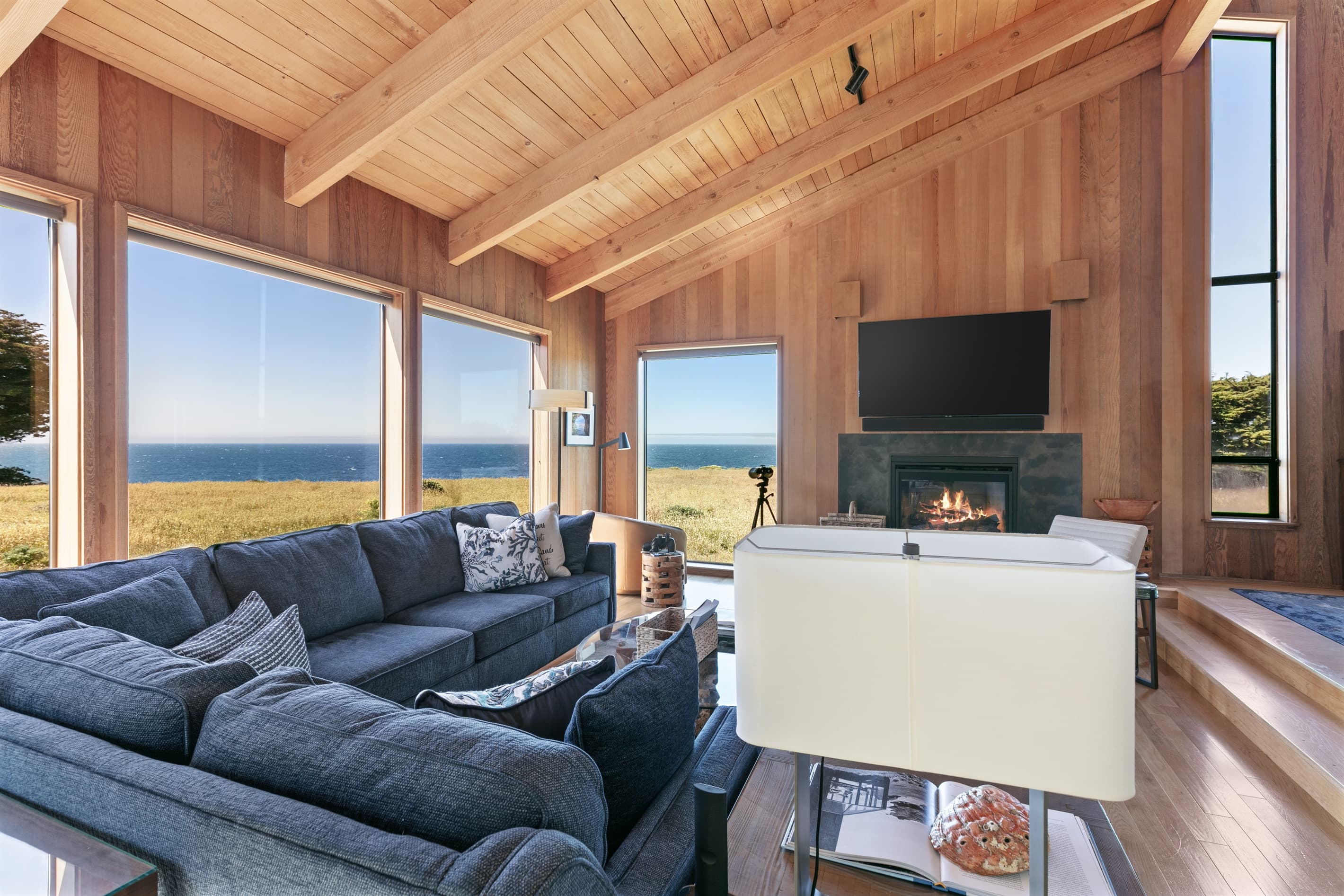 Expansive living room with vaulted wood ceilings, a cozy fireplace, and wall-to-wall ocean views—designed for refined relaxation with plush seating, natural textures, and serene coastal ambiance. 