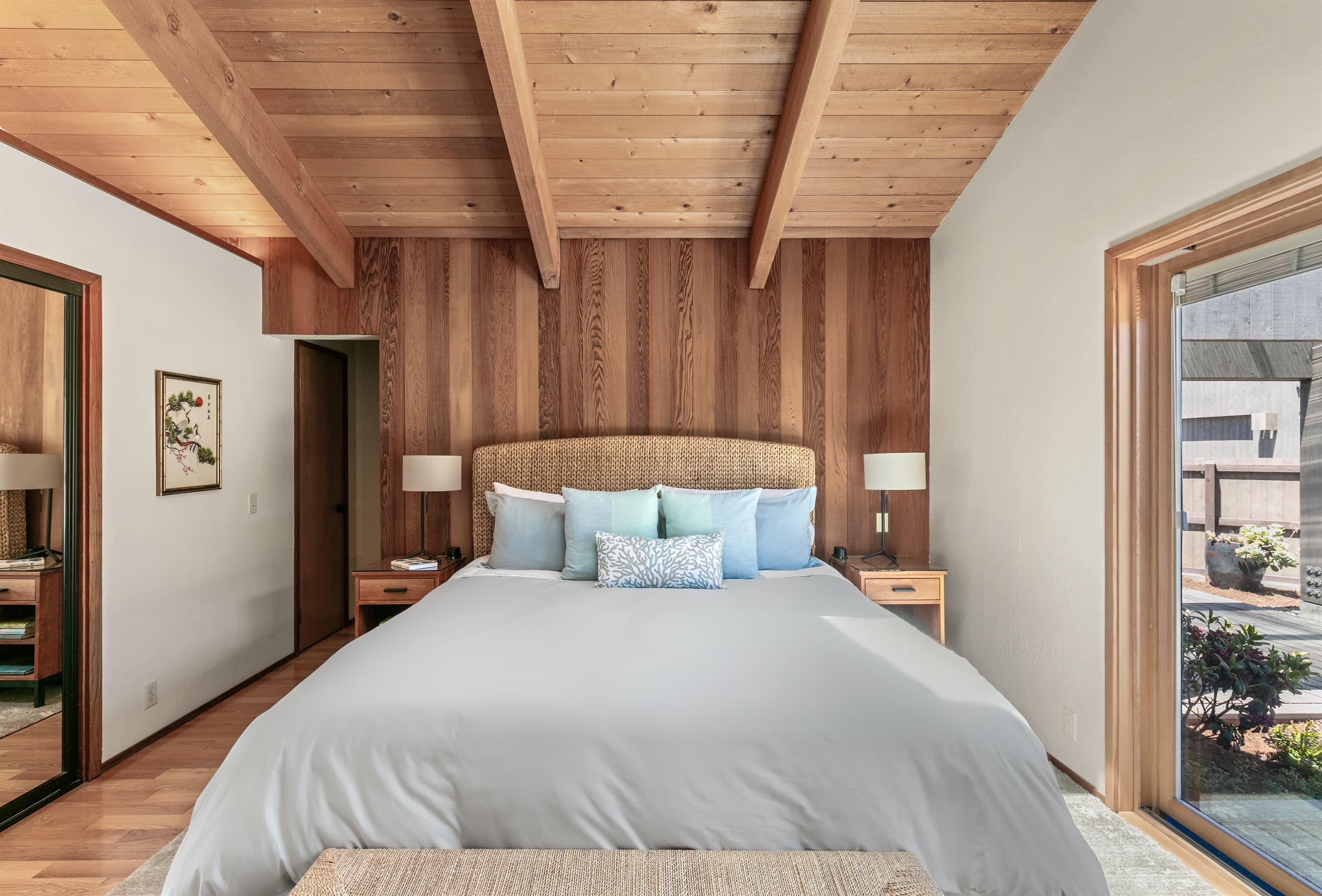 Serene primary bedroom features king-sized bed with woven headboard, soft coastal-inspired bedding, vaulted wood-beamed ceiling. Skylight has blackout blinds. Sliding glass door opens to a private courtyard. Ensuite bath at end of hall.