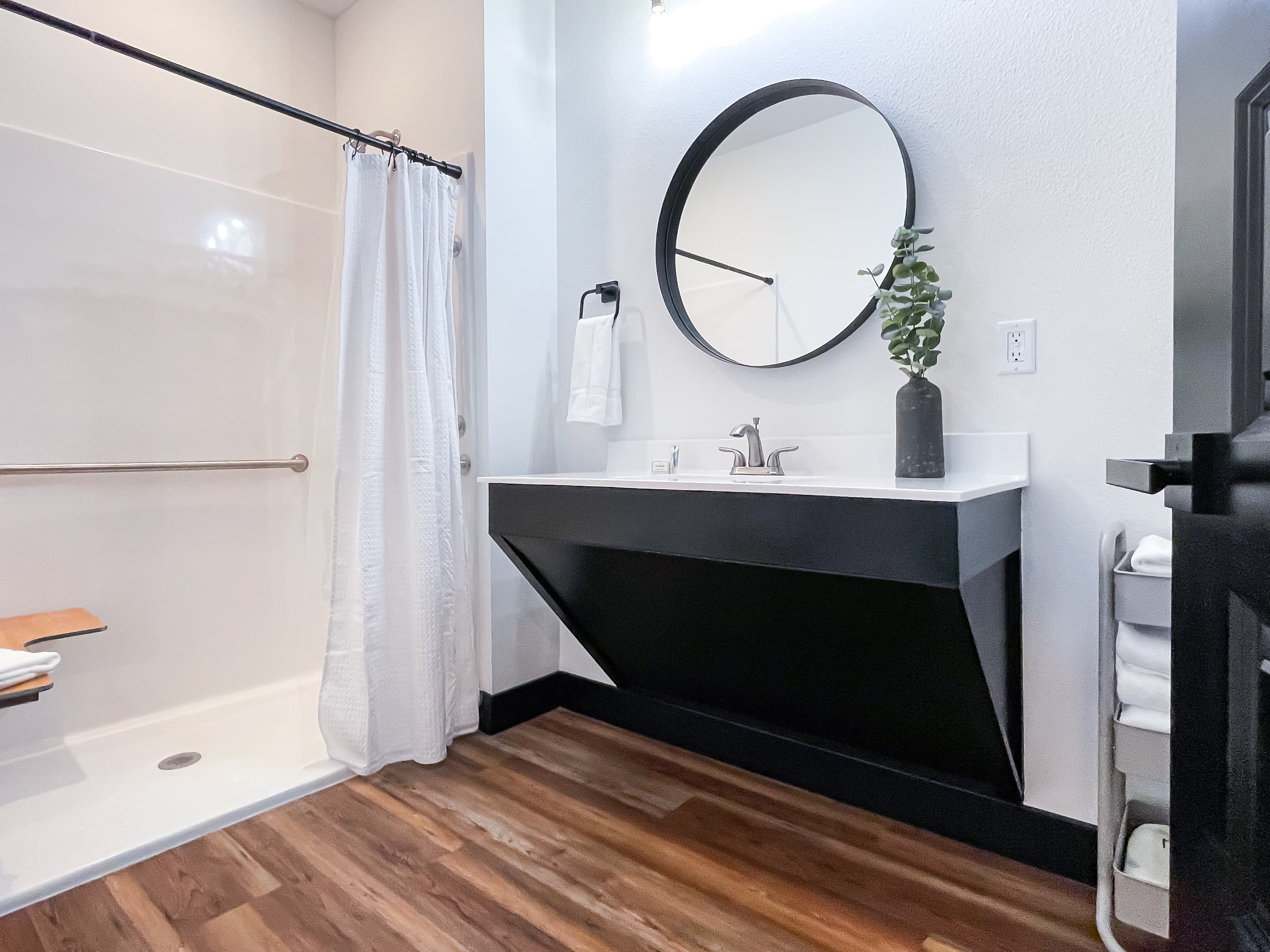 Main level en suite bathroom with a sloped vanity and grab bars on wall behind & next to toilet. The shower has a built in seat, grab bar and handheld shower head.