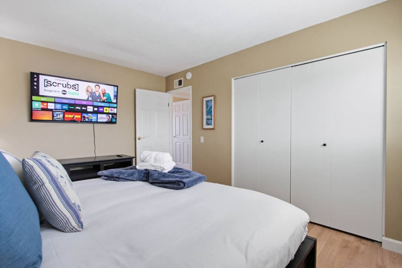 Bedroom w/ wall-mounted TV, wide closet & fresh towels set out on the bed.