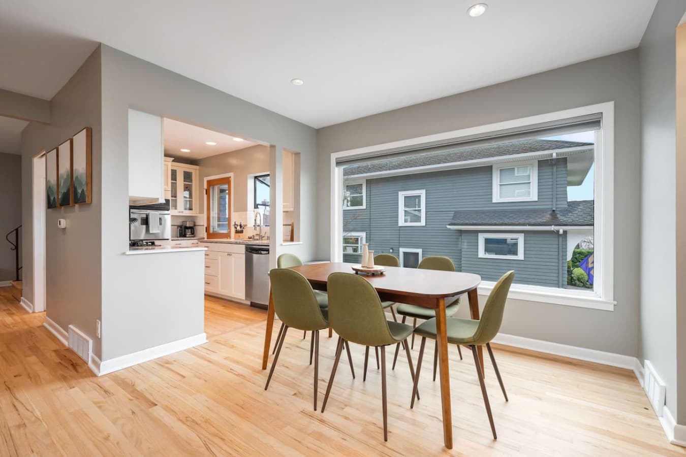 MAIN FLOOR- KITCHEN: A sleek wooden table with six olive-green chairs sits by a large window, filling the space with natural light. The open layout connects seamlessly to the kitchen with the dining room.