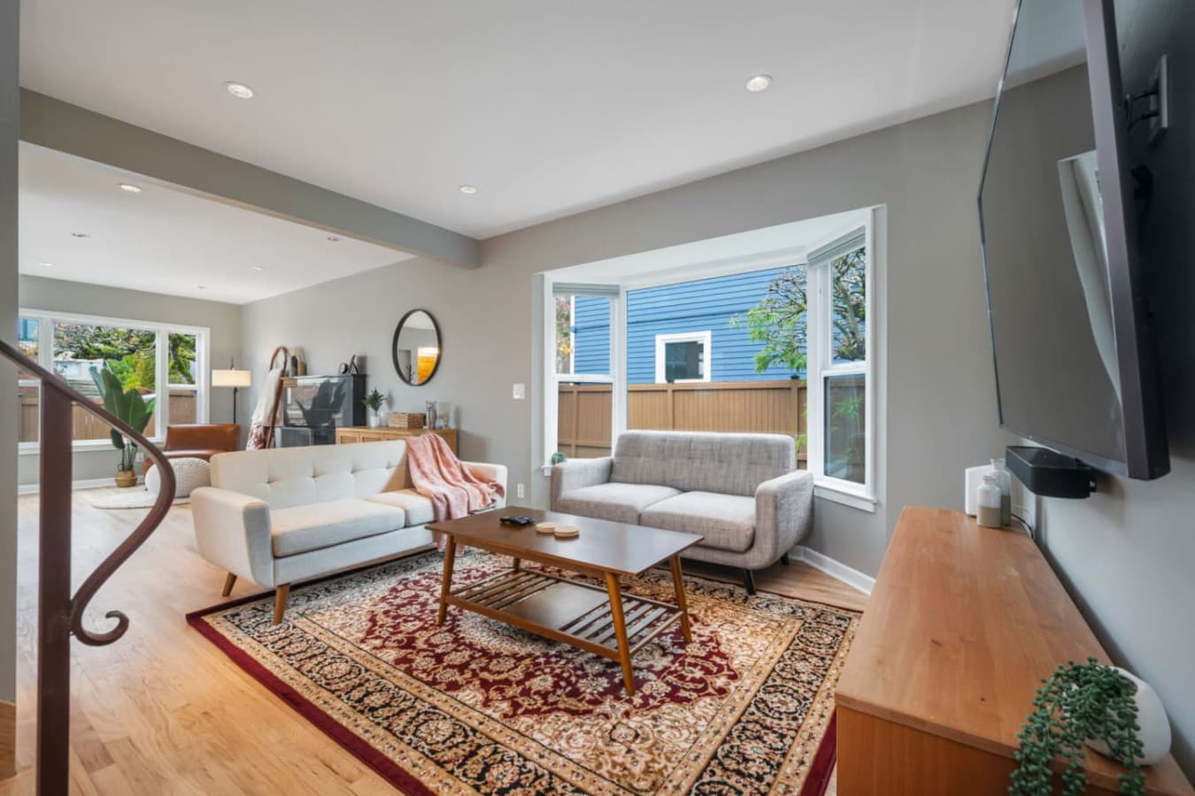 MAIN FLOOR - LIVING ROOM:  The inviting living room boasts large windows with lush views, a light gray sofa set for 2 people, a navy velvet love seat, and a Persian rug beneath a wooden coffee table.