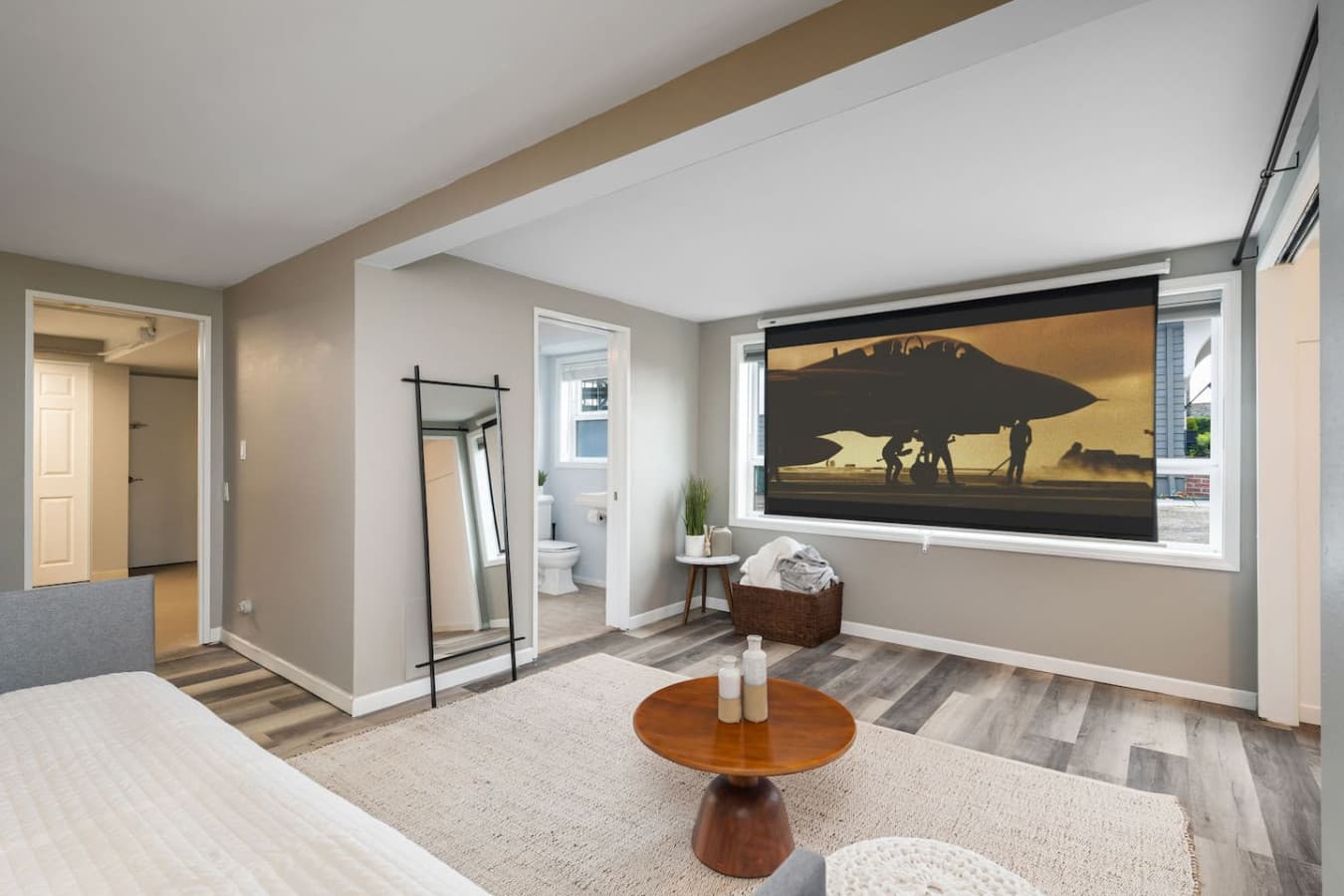 DOWNSTAIRS- BEDROOM: This modern and cozy bedroom features a neutral color palette, wood-like flooring, and a stylish minimalist design. It includes a comfortable bed, a wooden coffee table, a large rug, and a full-length mirror.