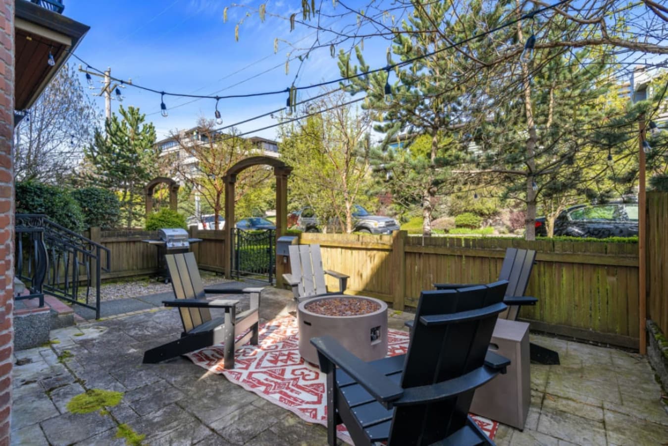 A cozy outdoor patio with Adirondack chairs around a fire pit, string lights, and a BBQ grill, perfect for relaxing and dining in a private, scenic setting.