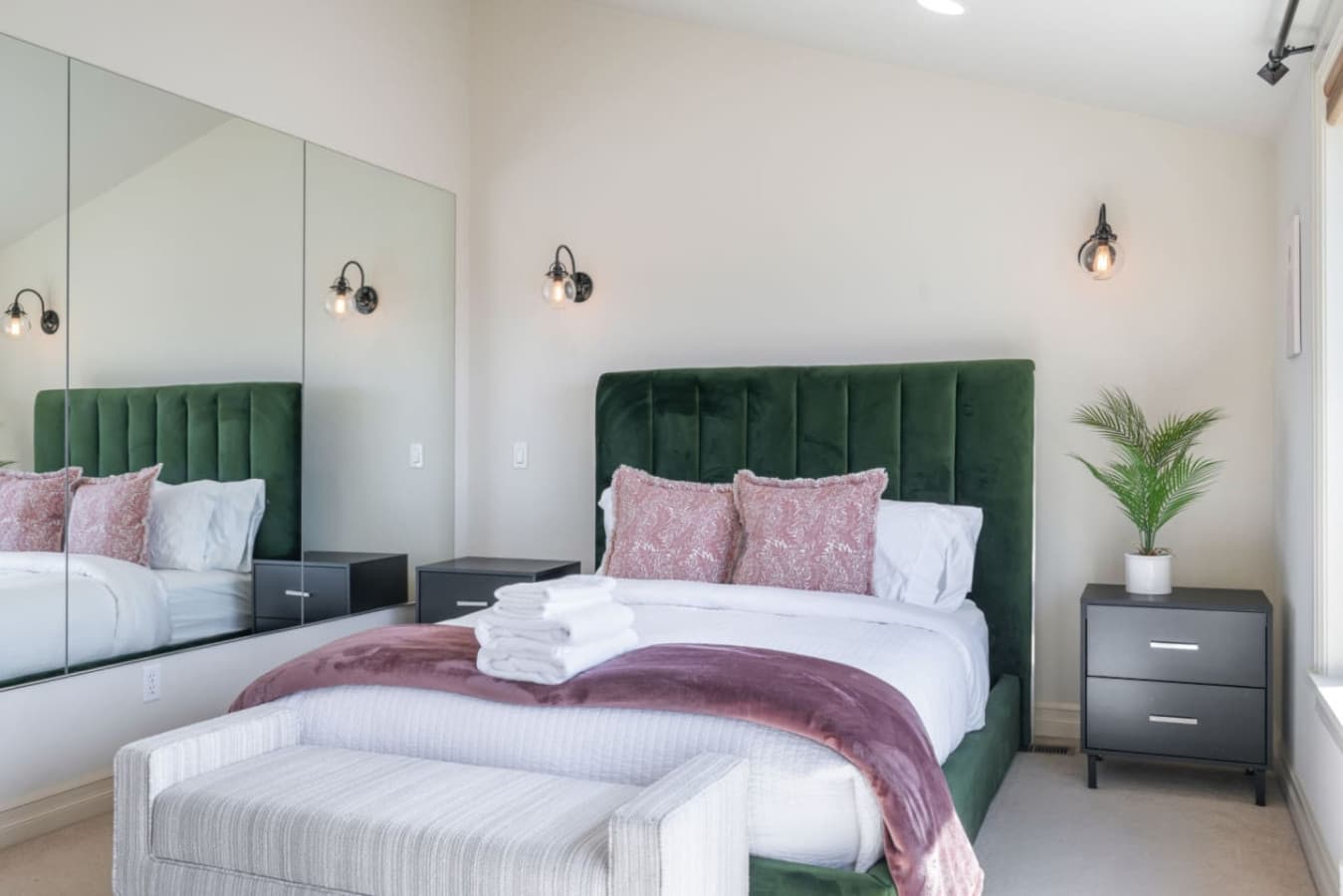 The bedroom features a large mirrored wall, which enhances the sense of space and reflects natural light, making the room feel brighter and more open. The mirror also adds a touch of sophistication to the modern decor, complementing the plush green v