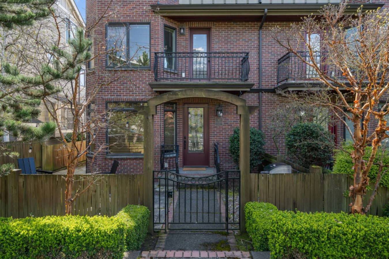 A welcoming entrance with a wooden pergola, wrought iron gate, and a short staircase leading to a brick façade with a glass-paneled door, blending natural elements with classic elegance.
