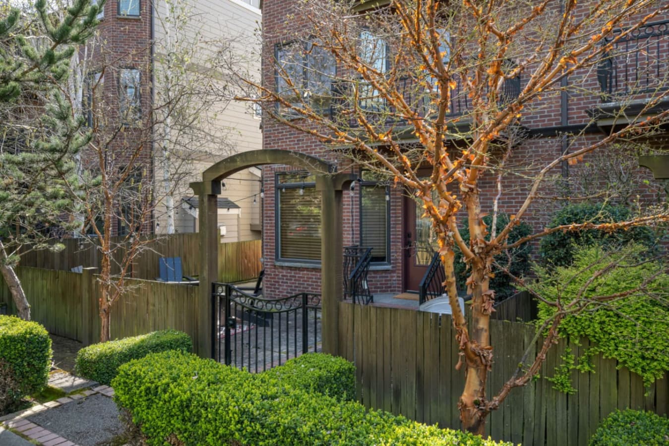 A welcoming entrance with a wooden pergola, wrought iron gate, and a short staircase leading to a brick façade with a glass-paneled door, blending natural elements with classic elegance.