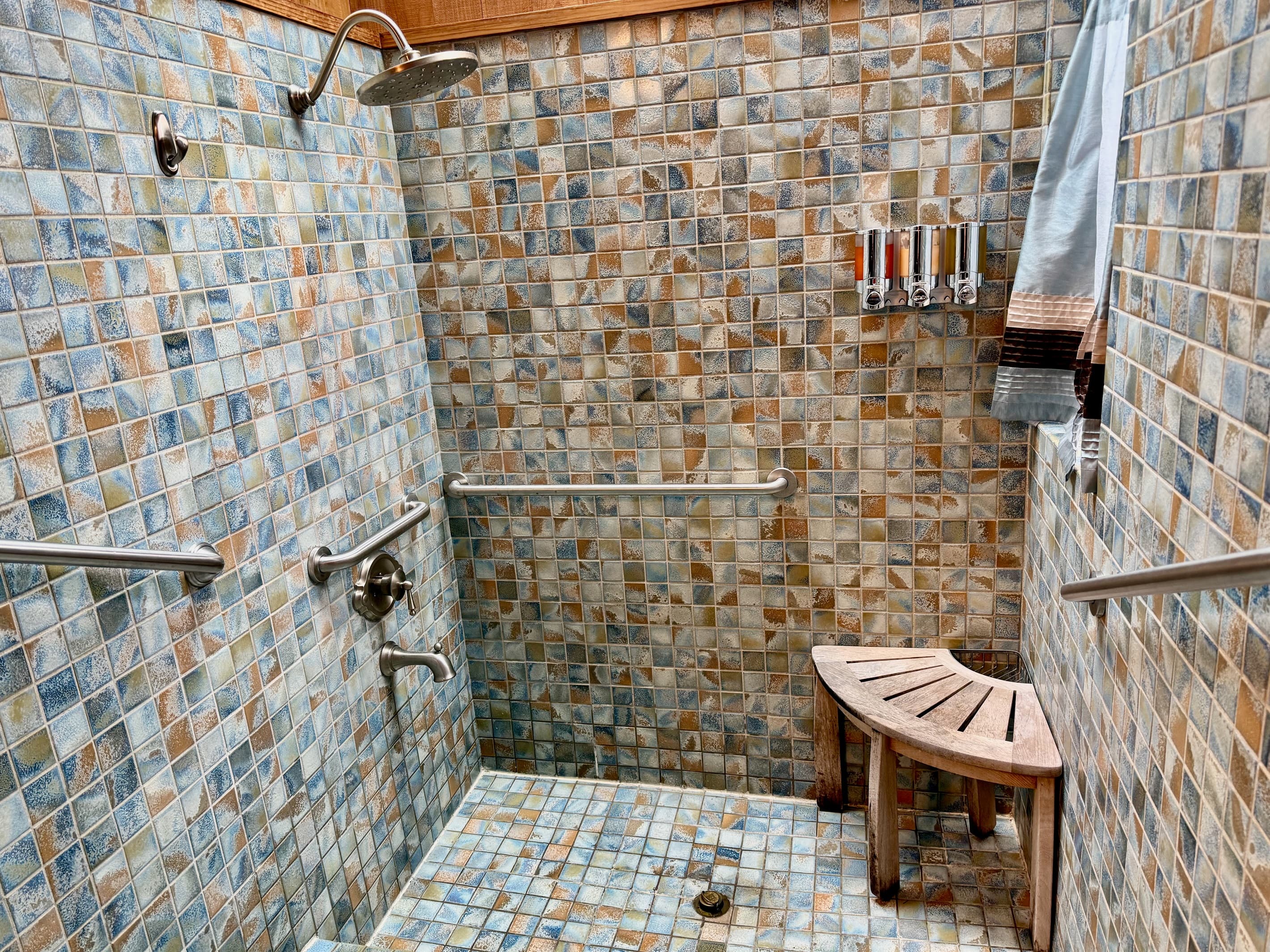 Elegant walk-in tiled Roman sunken shower (3-steps), a soothing rain shower head, and a teak corner bench. Framed by a skylight and natural wood paneling, this serene retreat offers spa-like indulgence with premium soap and shampoo dispensers.