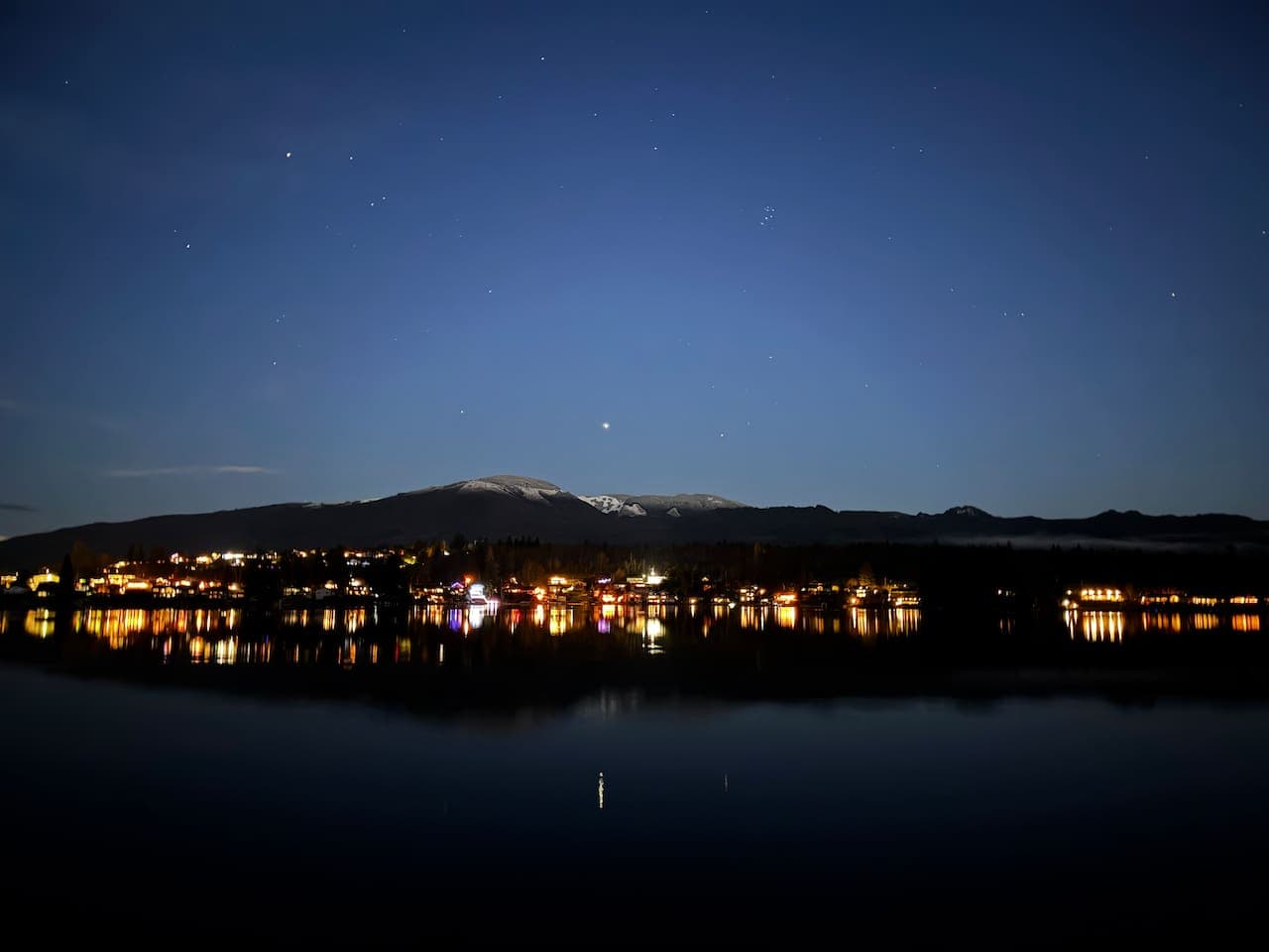 Lights from the far shoreline shimmer across the lake as the deep blue sky fades toward the horizon. The water reflects the quiet glow, creating a peaceful evening scene that feels calm, open, and still.