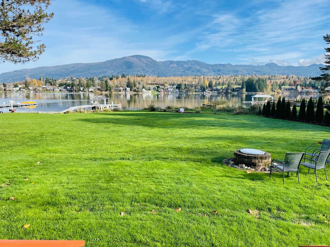 A wide, green backyard stretches to the lake, with a dock and fire pit ready for evenings outdoors. The open lawn feels inviting, and the calm water reflects the shoreline, creating a peaceful connection between land and lake.