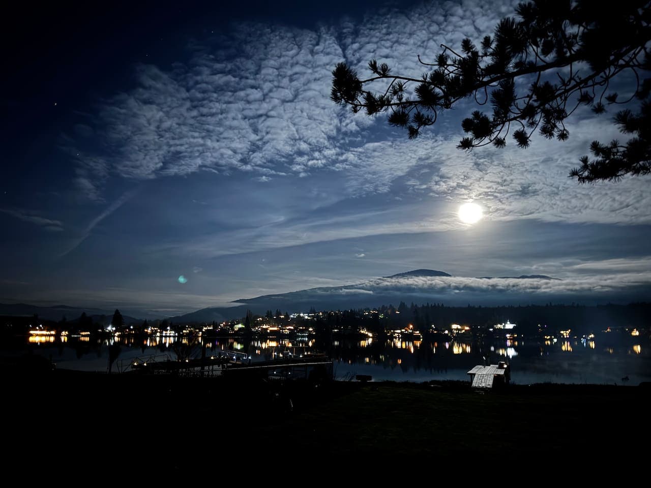 A glowing moon rises above a clouded sky, casting light over the lake as shoreline lights shimmer on gentle ripples. The horizon softens into a pale blue, creating a serene, moody evening where water, clouds, and moonlight meet.