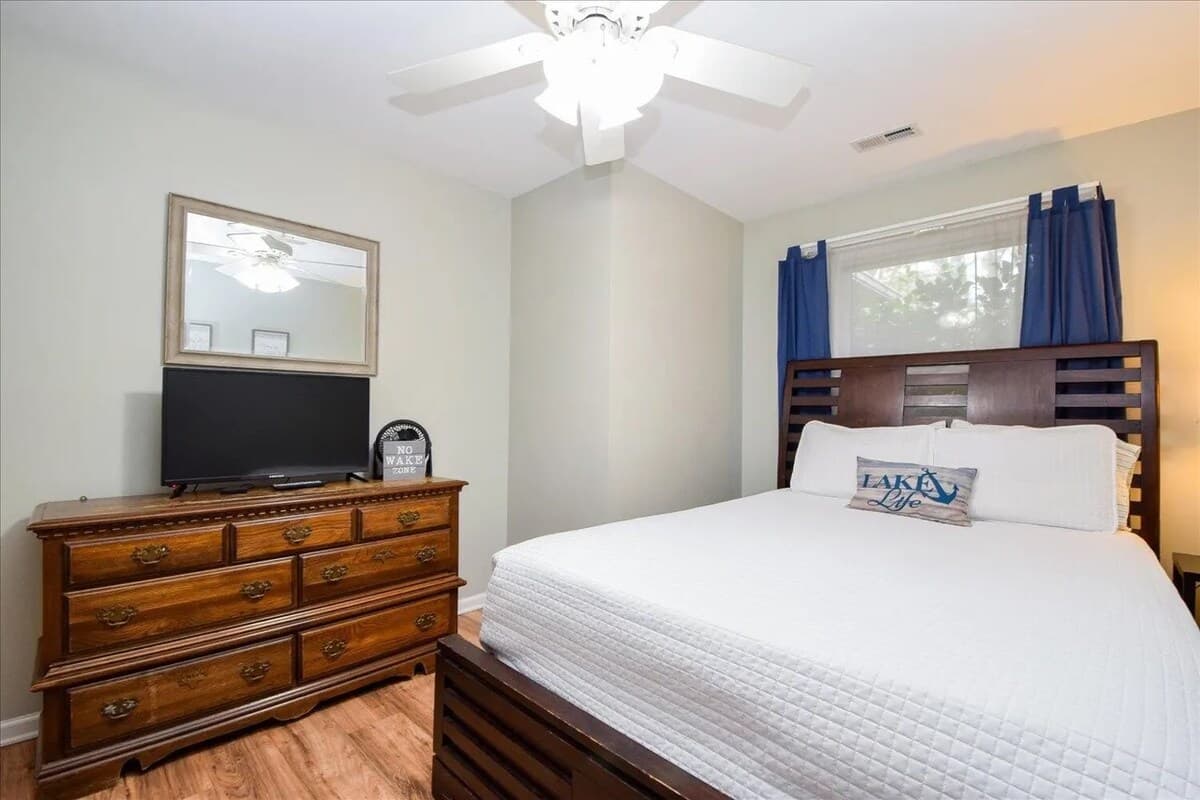 Sun-drenched queen guest room with honey-wood bed, navy curtains, and extra sleeper seating. Queen bed, lake-themed decor, flat-screen TV, gray walls, coastal accent pillow, driftwood flooring, Smith Mountain Lake getaway.