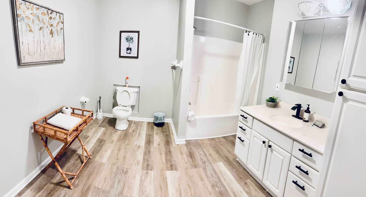 Roomy, light-filled bath — unwind after a day on the water.full bathroom, soaking tub, shower combo, rattan accent, luxury vinyl plank, boutique-hotel lighting, botanical art, lake house bathroom. 