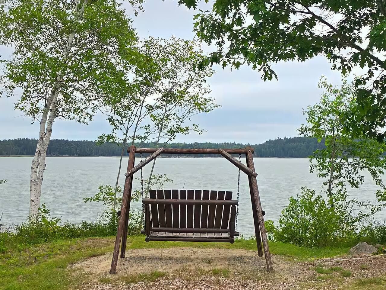 A rustic wooden swing faces the calm waters of the bay, framed by leafy trees and soft grass. It’s the perfect spot to sit back, sway gently, and take in the peaceful rhythm of nature.