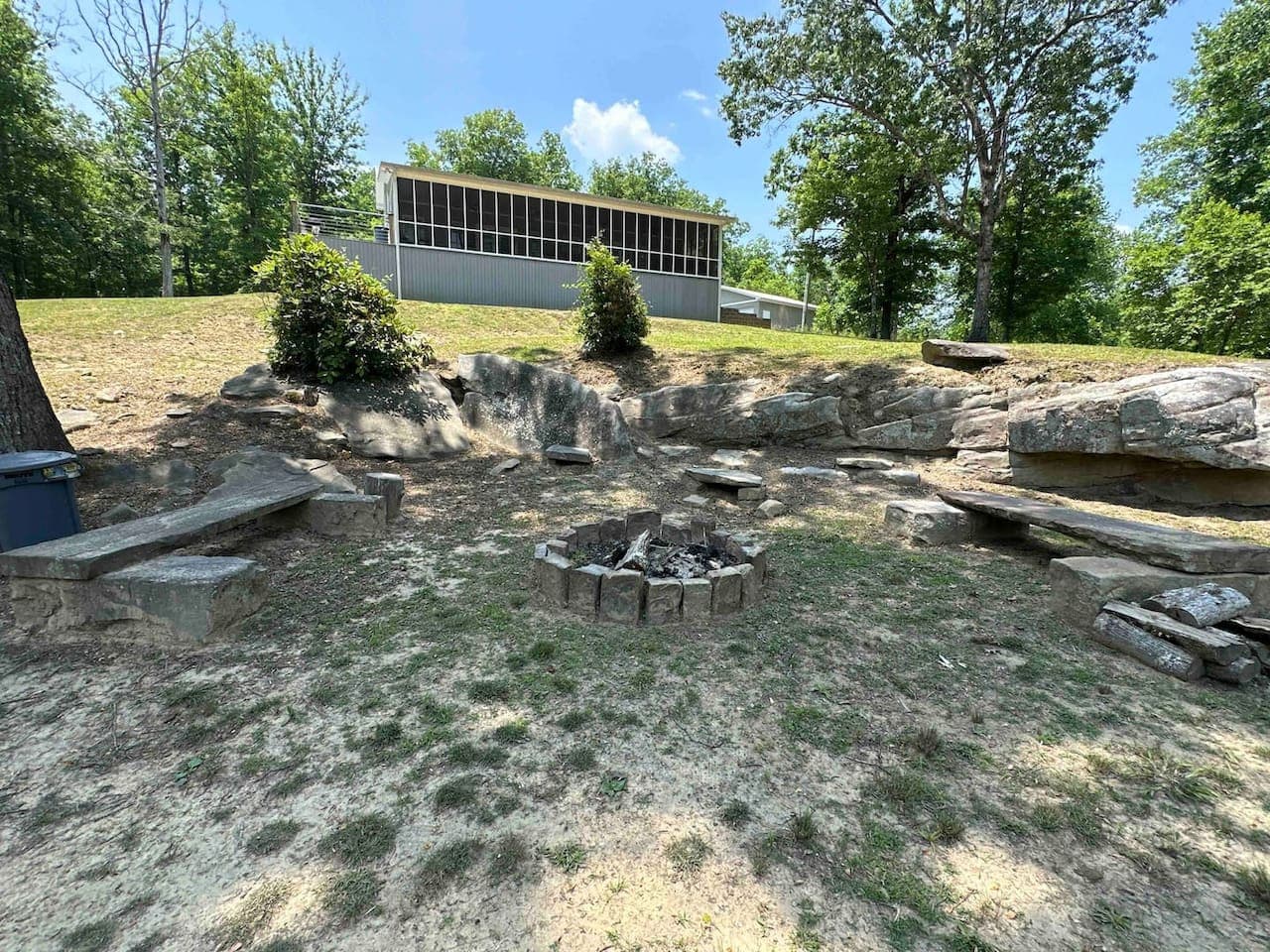Backyard outdoor fire pit area with stone seating and a natural rock setting, surrounded by trees and overlooking the house.