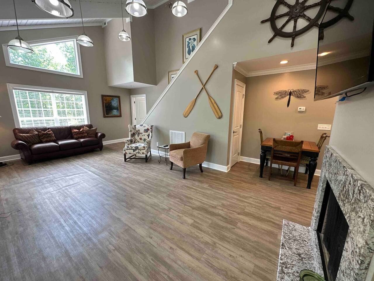 Bright, open-concept living and dining area with vaulted ceilings, large windows, wood-style floors, and neutral decor. Features a leather sofa, accent chairs, nautical wall art with oars and a ship wheel, a wooden dining set, and a stone fireplace.