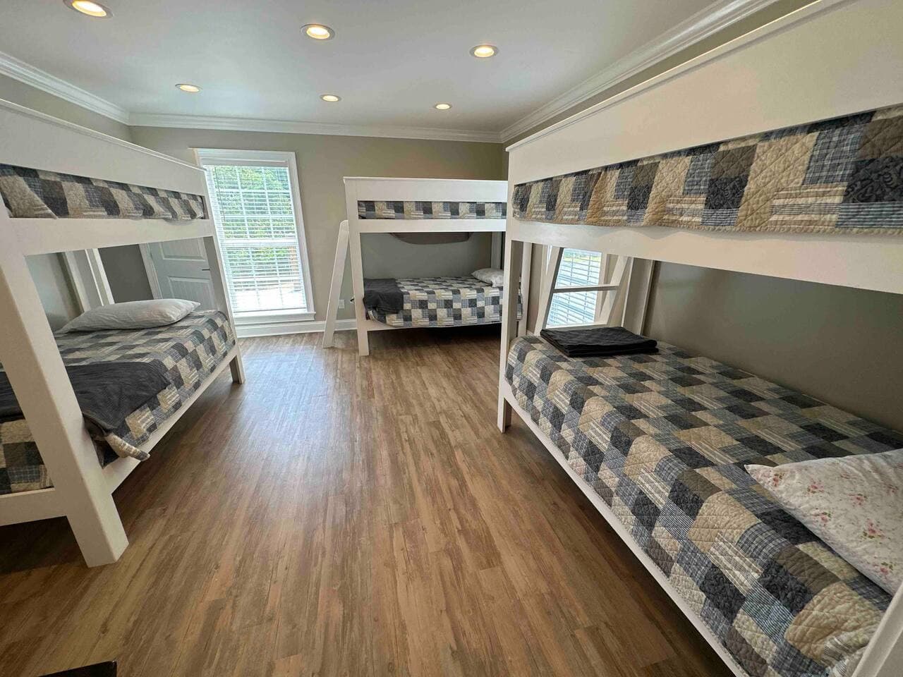 Bright bedroom with three white wooden bunk beds, checkered bedding in gray and blue tones, hardwood floors, recessed lighting, and large windows letting in natural light.