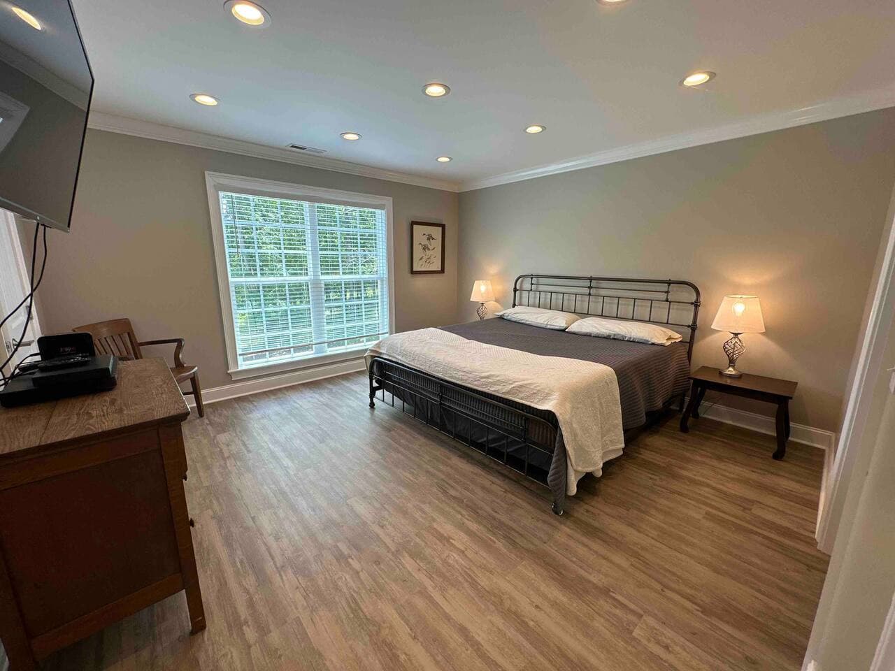Spacious bedroom with a king bed, hardwood floors, and large windows offering plenty of natural light. Features include a mounted TV, dresser, and cozy bedside lighting for a relaxing stay.