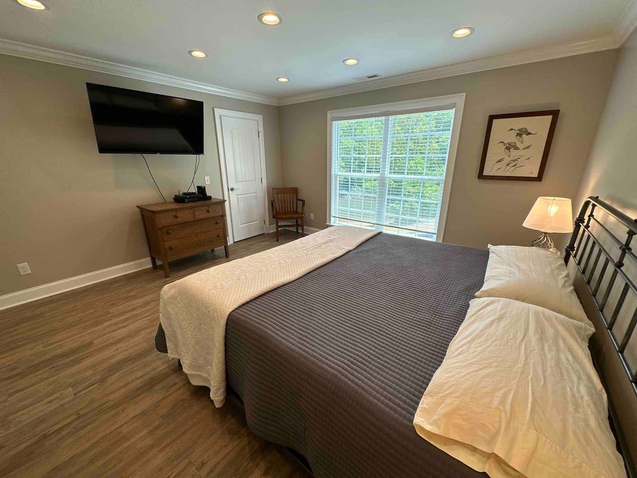 Bright and inviting room with a king-size bed, modern lighting, and elegant wood flooring. Includes a TV, dresser, and large window with peaceful outdoor views.