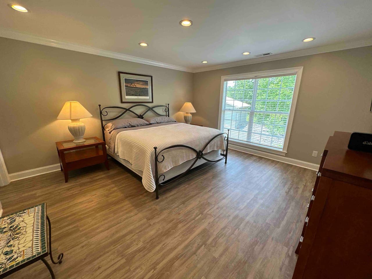 Spacious bedroom with a large window allowing ample natural light, featuring a queen bed with a decorative metal frame, wooden nightstands with lamps, and neutral tones for a calm, comfortable atmosphere.