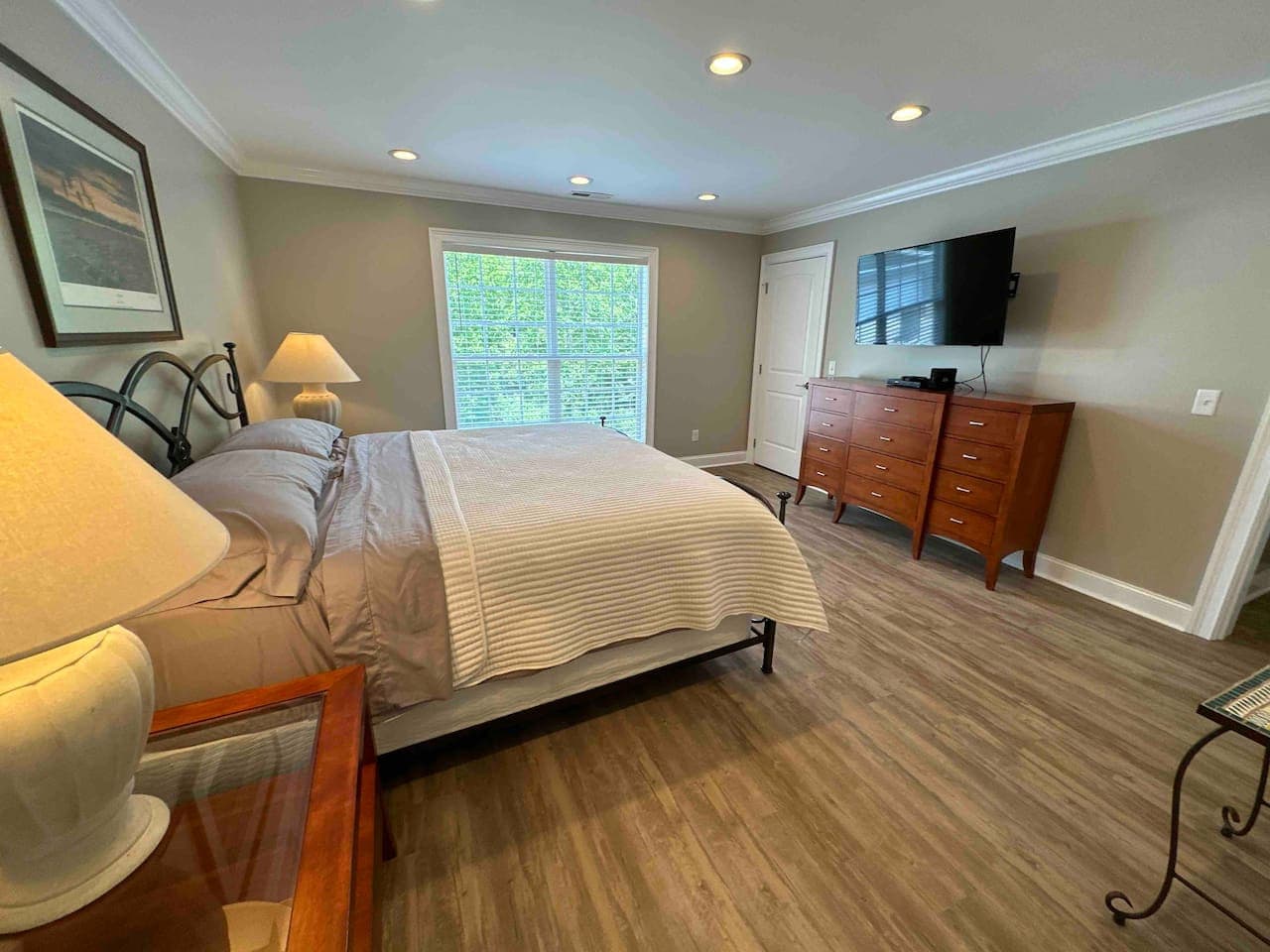 Bright and elegant bedroom with wood flooring, soft lighting, and a cozy bed centered against a light-colored wall. The large window offers serene outdoor views, creating a peaceful retreat.