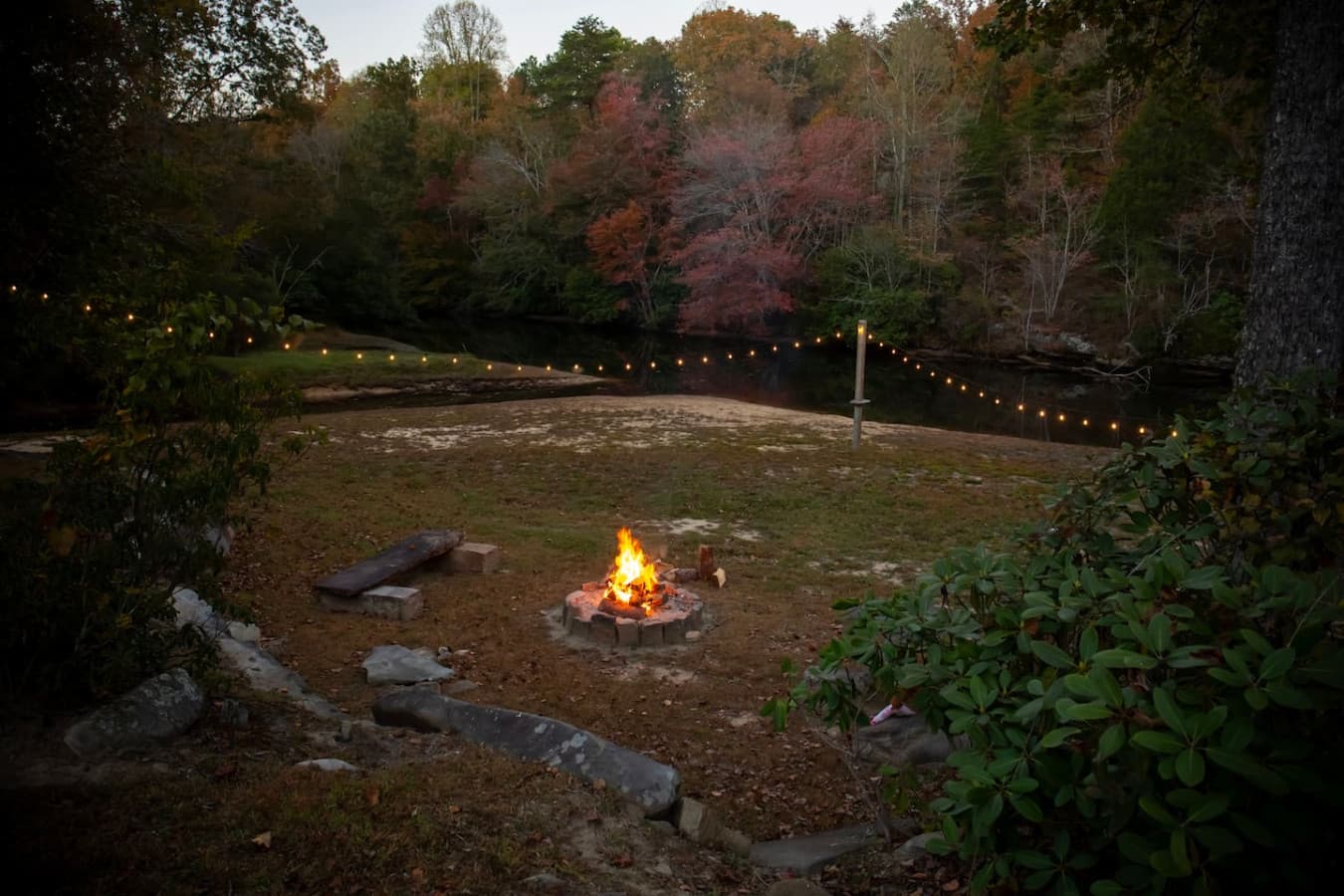 Scenic outdoor fire pit area by the water, surrounded by string lights and natural landscaping, creating a cozy evening atmosphere.
