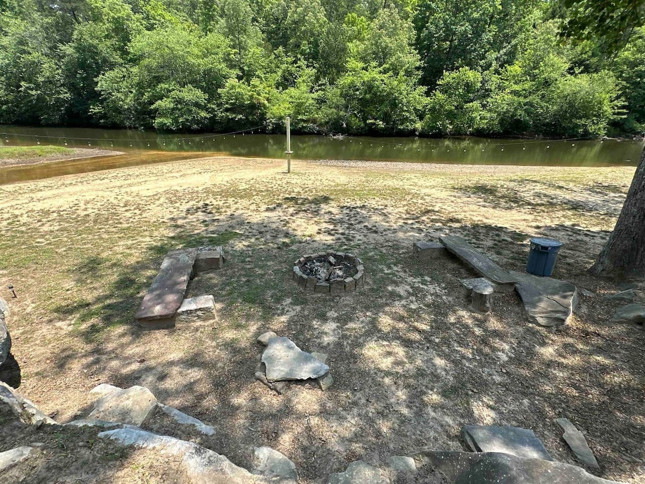 Riverside fire pit area with rustic stone and wooden seating, offering a peaceful outdoor spot surrounded by trees and natural scenery.