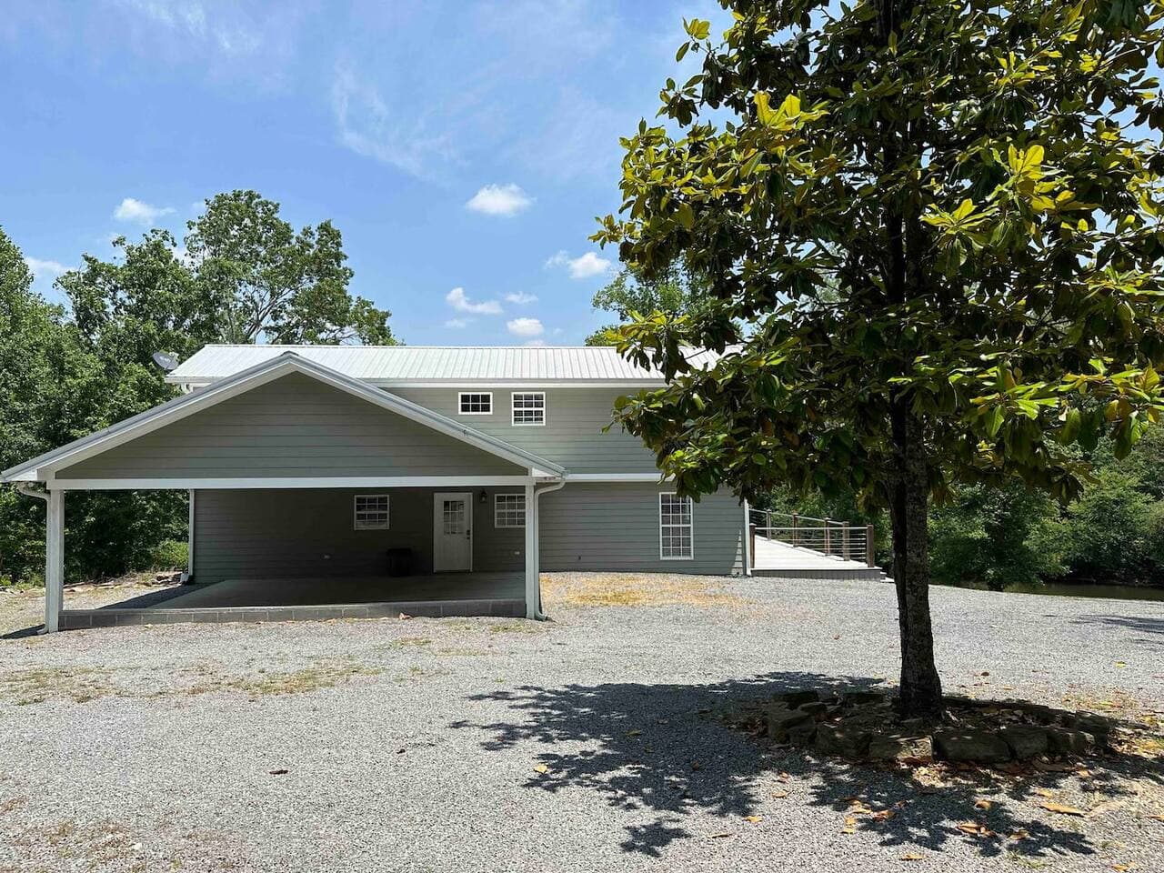 Front exterior of the home featuring a covered carport, gravel driveway, and mature tree providing shade and curb appeal.