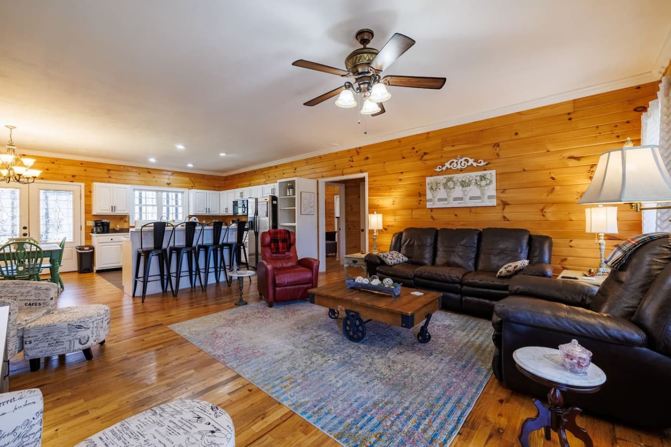 Cozy cabin living room with warm wood walls, leather sofas, a rustic coffee table, and an open kitchen perfect for gatherings.