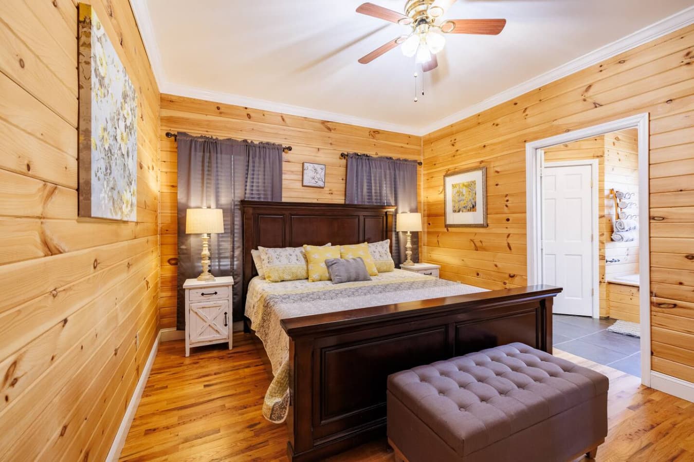 Bright and welcoming king bedroom featuring rich wood furnishings, cozy lighting, and soft yellow accents. The en-suite bath and tufted bench add a touch of comfort and luxury—perfect for a peaceful cabin retreat.