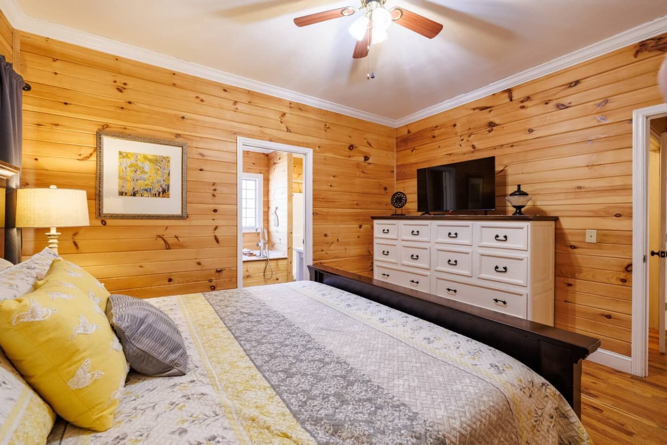 Sunny and stylish king bedroom with golden wood walls, plush bedding, and a big-screen TV—perfect for cozy mornings, movie nights, or just kicking back in cabin comfort.