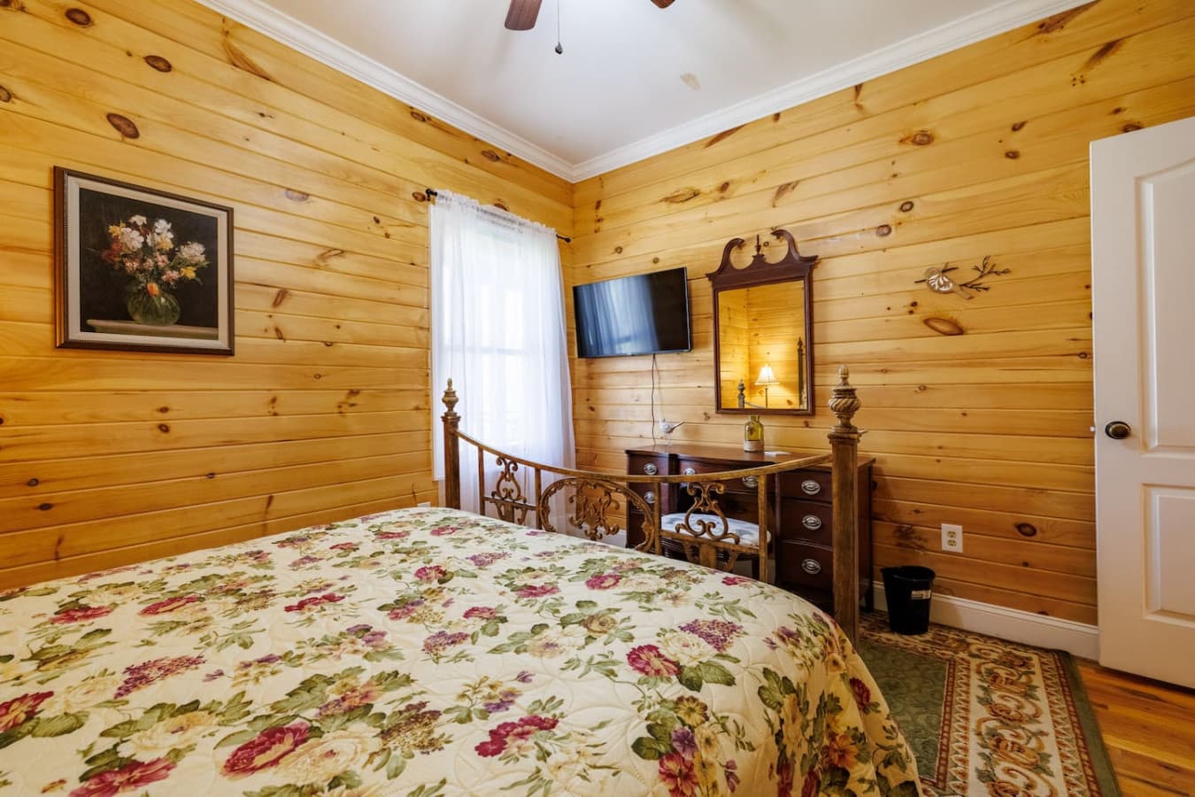 Queen bedroom with warm wood walls, a vintage-style iron bed dressed in floral linens, and a cozy setup featuring a wall-mounted TV and antique vanity—perfect for a restful, nostalgic retreat.