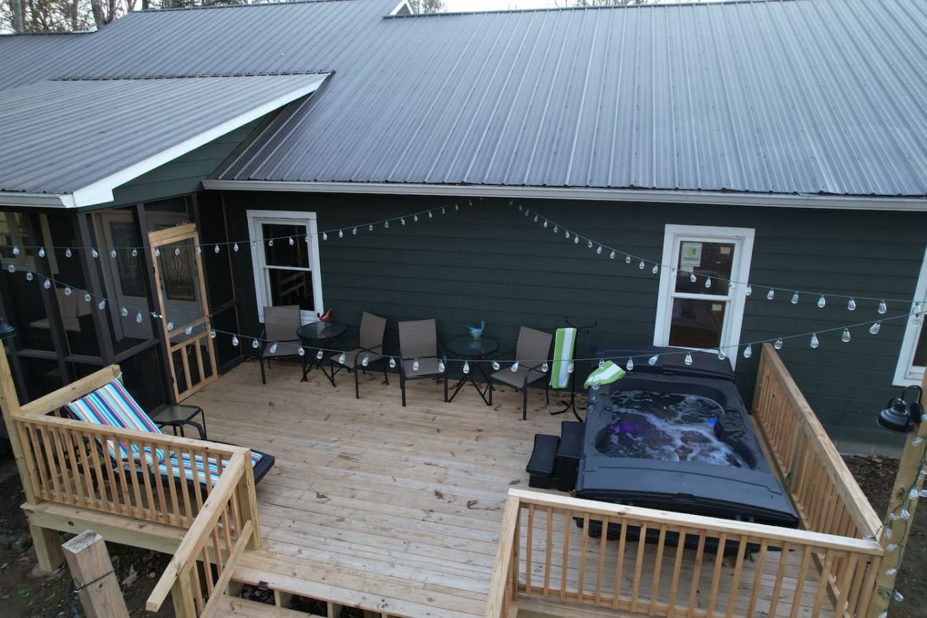 Enjoy the spacious back deck at The Bird House, featuring a relaxing hot tub, comfortable outdoor seating, and charming string lights, all set against a peaceful, wooded backdrop for the perfect private retreat.