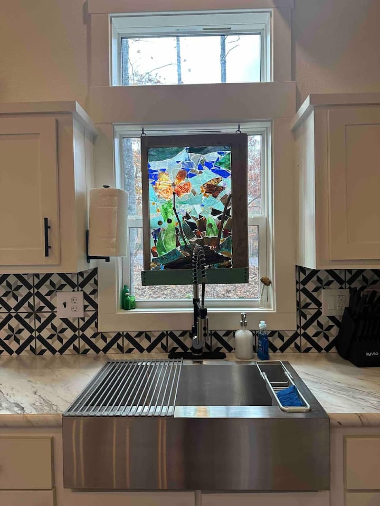 The kitchen sink sits beneath a window adorned with colorful mosaic art, adding a bright and artistic touch to your cooking space.