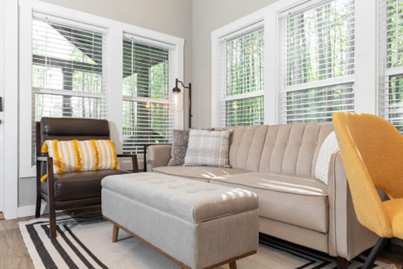 Bright and modern living room featuring a comfy sofa, accent chair, and ottoman, surrounded by large windows that fill the space with natural light.