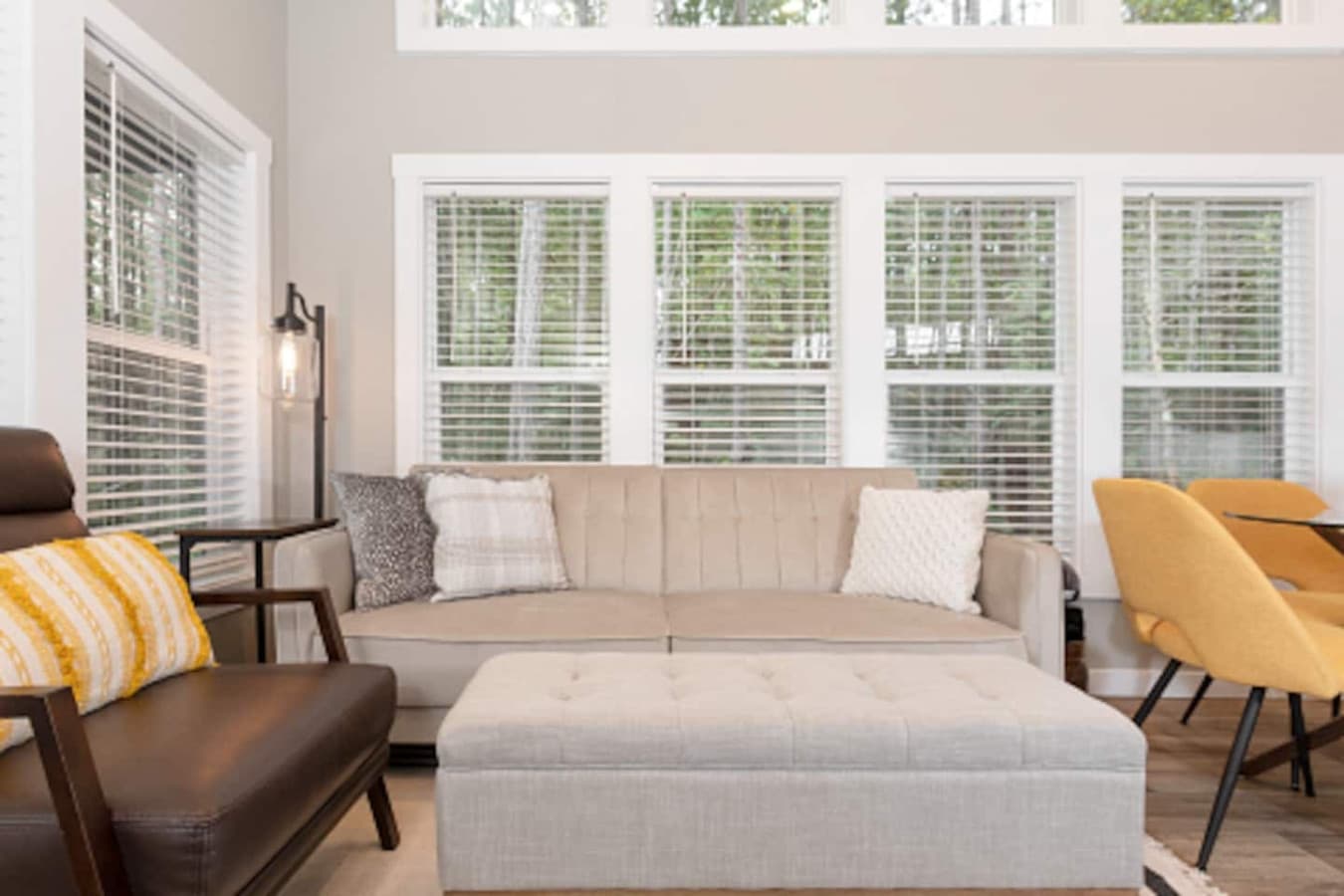 Bright and modern living room featuring a comfy sofa, accent chair, and ottoman, surrounded by large windows that fill the space with natural light.
