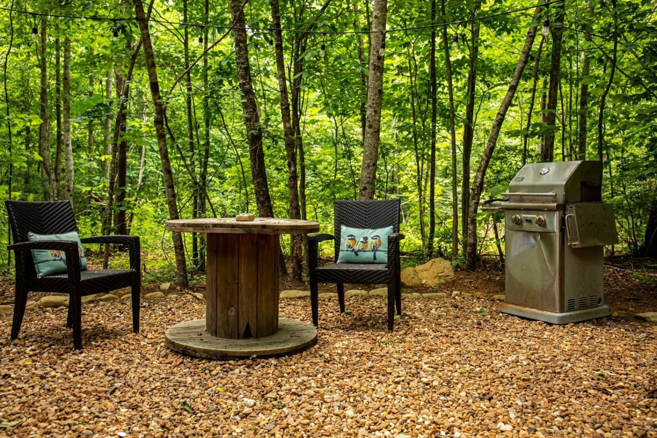 The gravel patio behind the home features a propane grill, string lights, and a cozy sitting area. The trail to the creek starts just to the right.