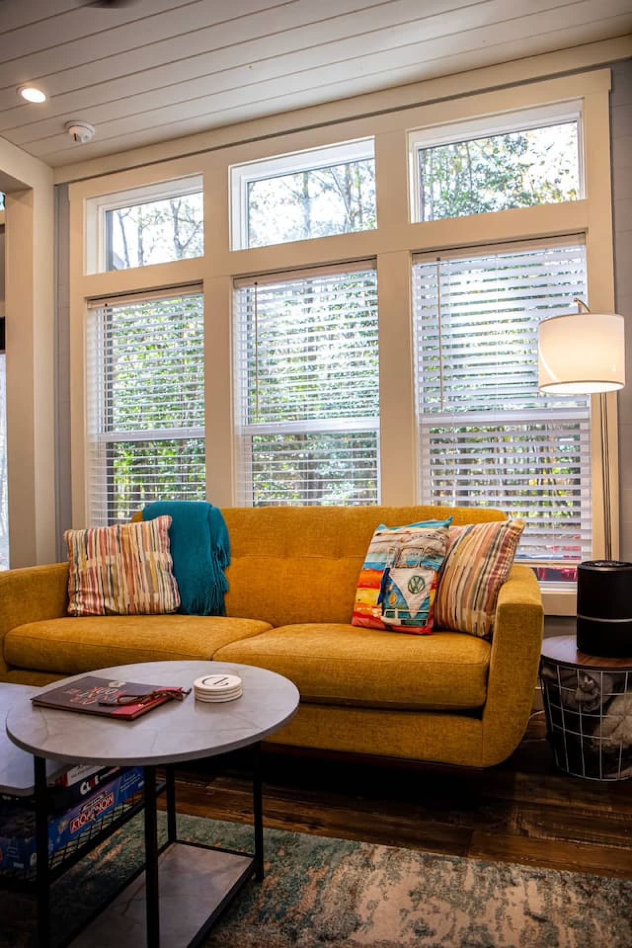 The cozy yellow sofa, colorful pillows, and soft lighting create a warm ambiance, while the large windows bring the outdoors in, offering a peaceful view of the trees outside.