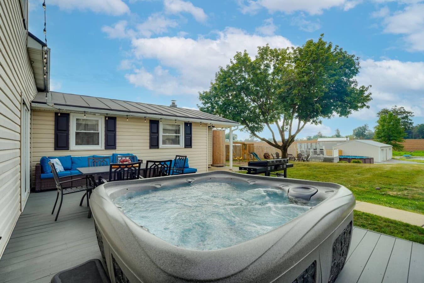 A peaceful suburban patio with a dining table, hot tub, and a shed, surrounded by greenery.