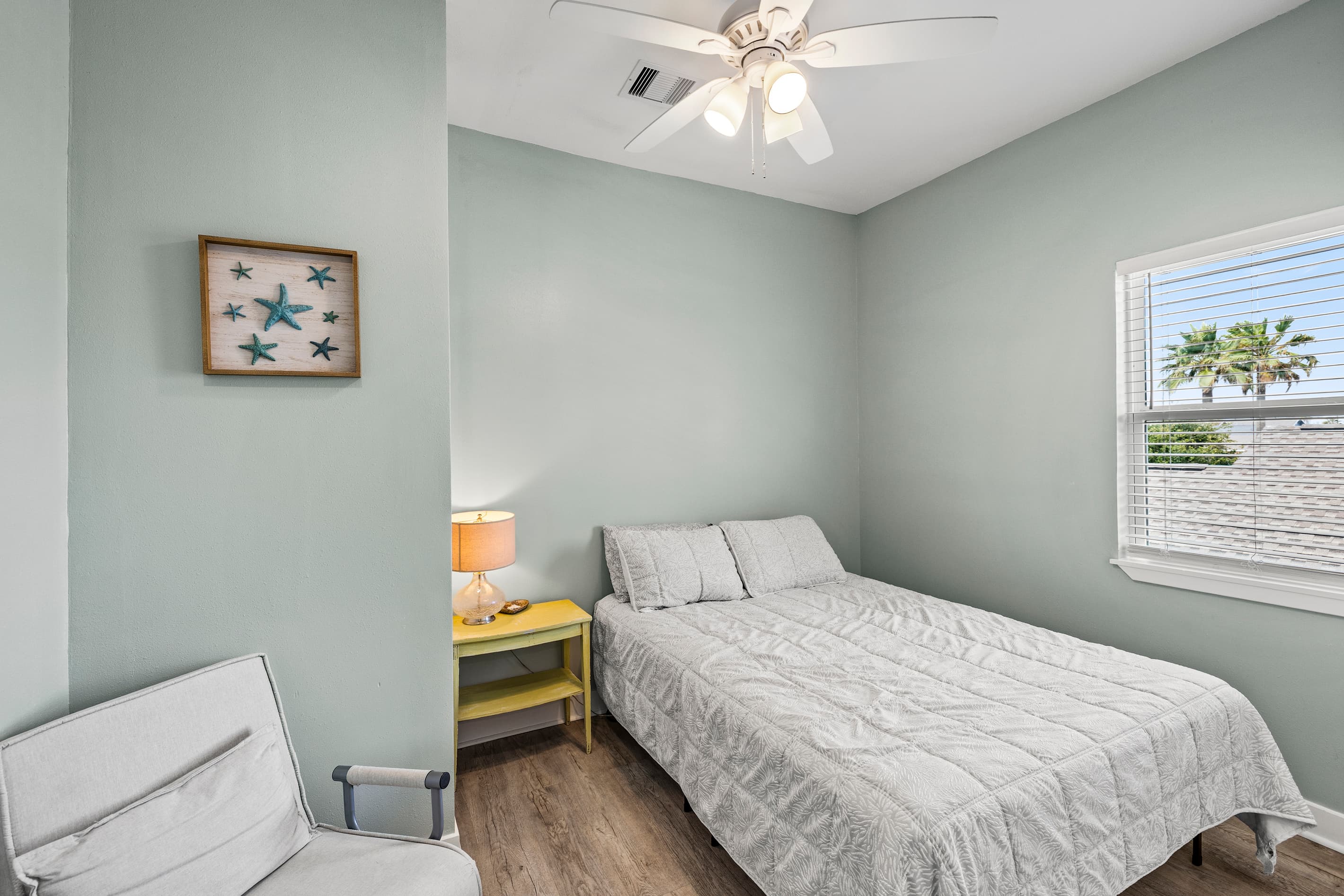 Bedroom 1 at The Lookout at R½ — a queen bed, coastal starfish art, and a window framing a Galveston palm tree. Wake up to island vibes every morning.