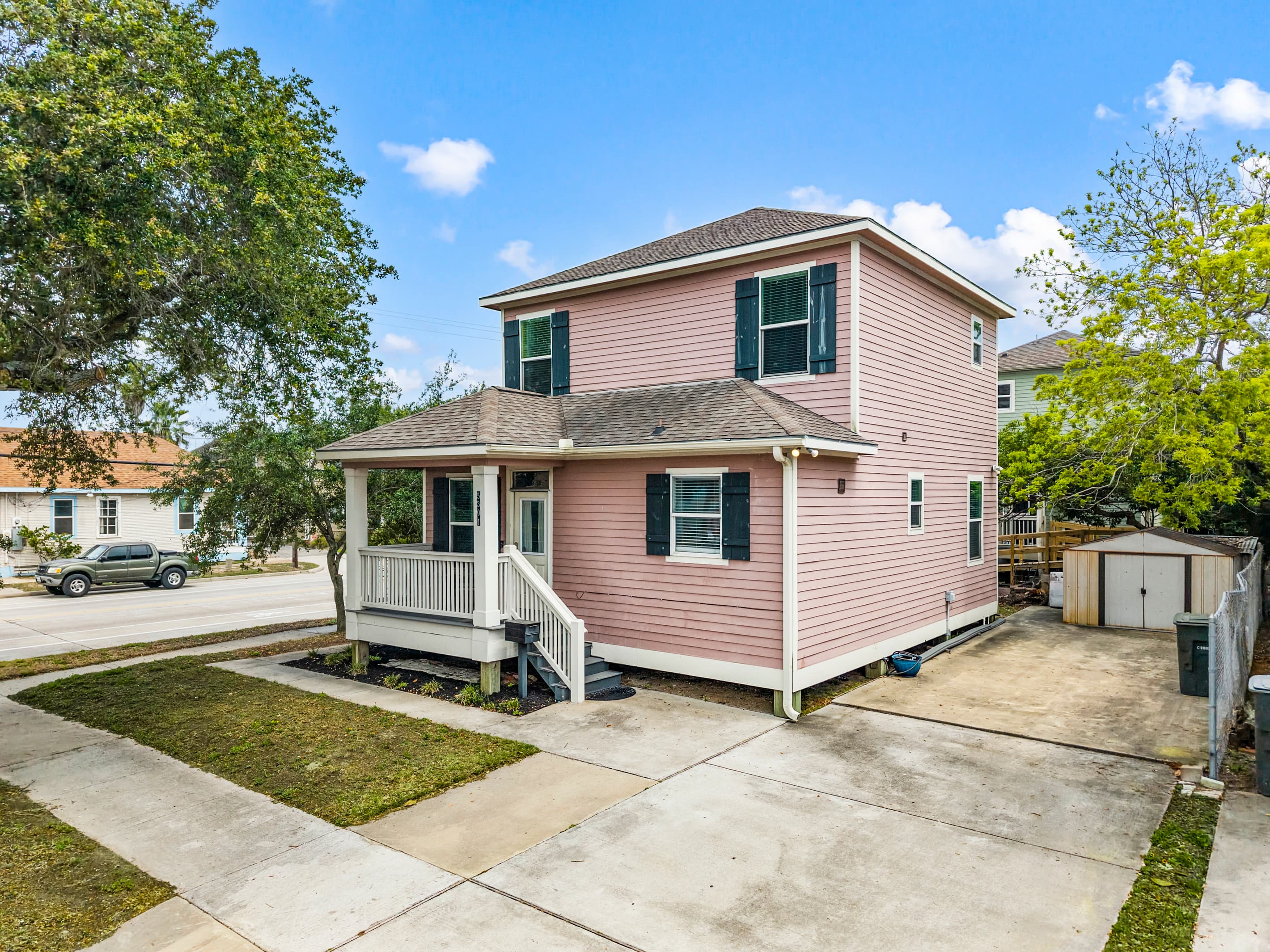 Summer Bliss from the street — the full corner home with a large driveway, lush lawn, and a storage shed. A well-maintained Galveston property steps from the beach.