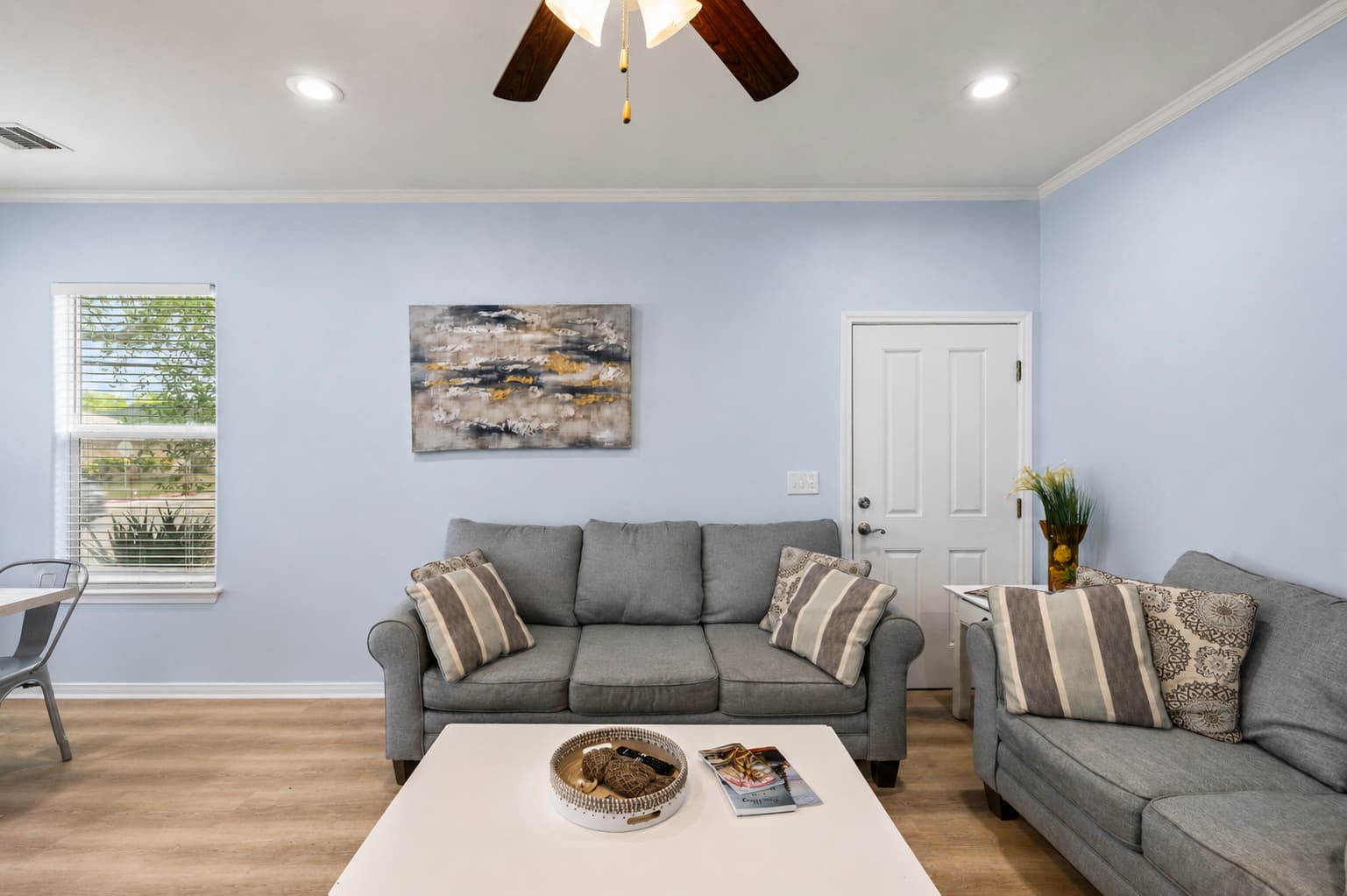 The living room at Summer Bliss — a plush gray sofa, white coffee table, and coastal wall art. Light blue walls and hardwood floors make it feel like a home away from home.