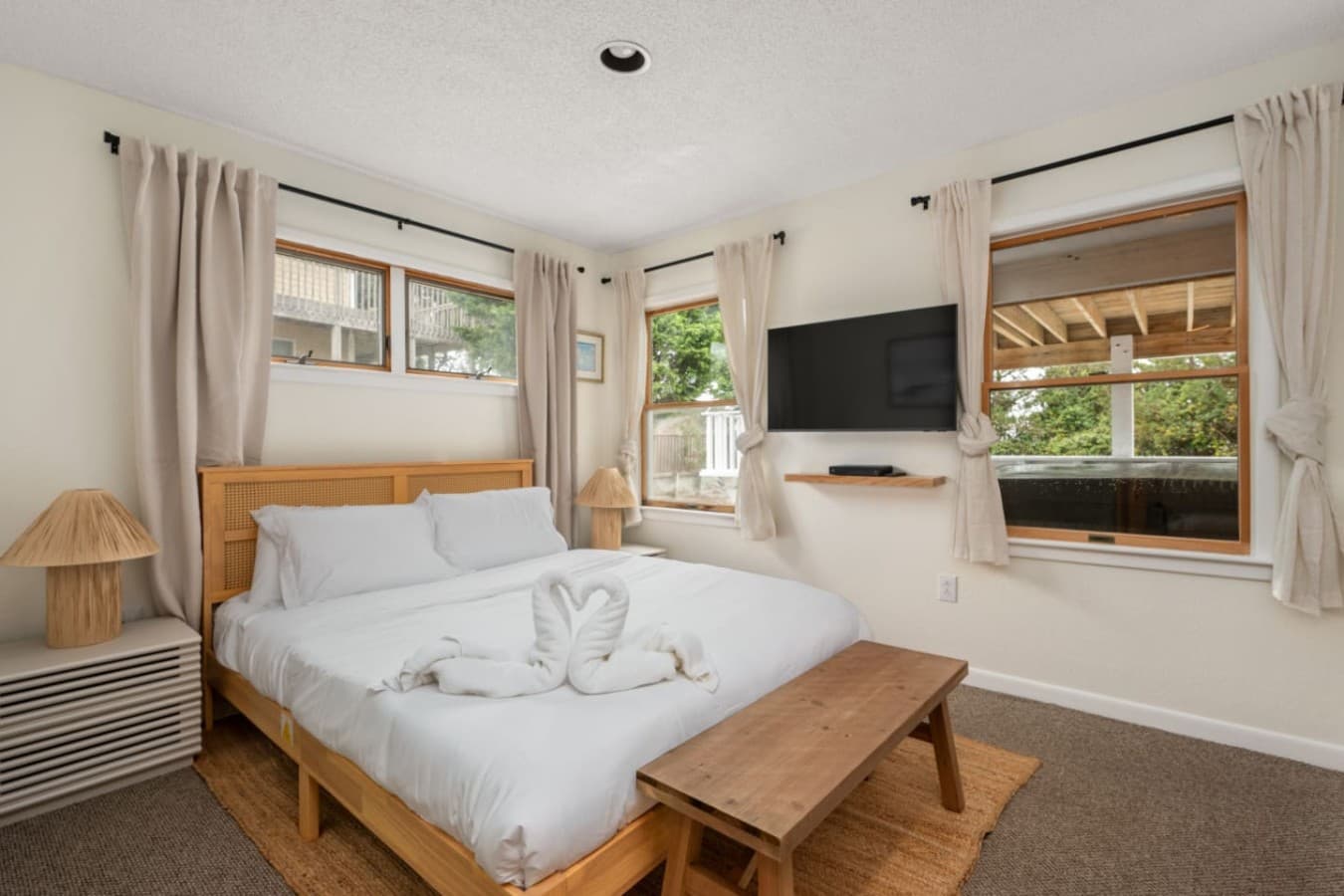 Sunlit bedroom w/ plush bedding, TV & leafy views through wide windows.