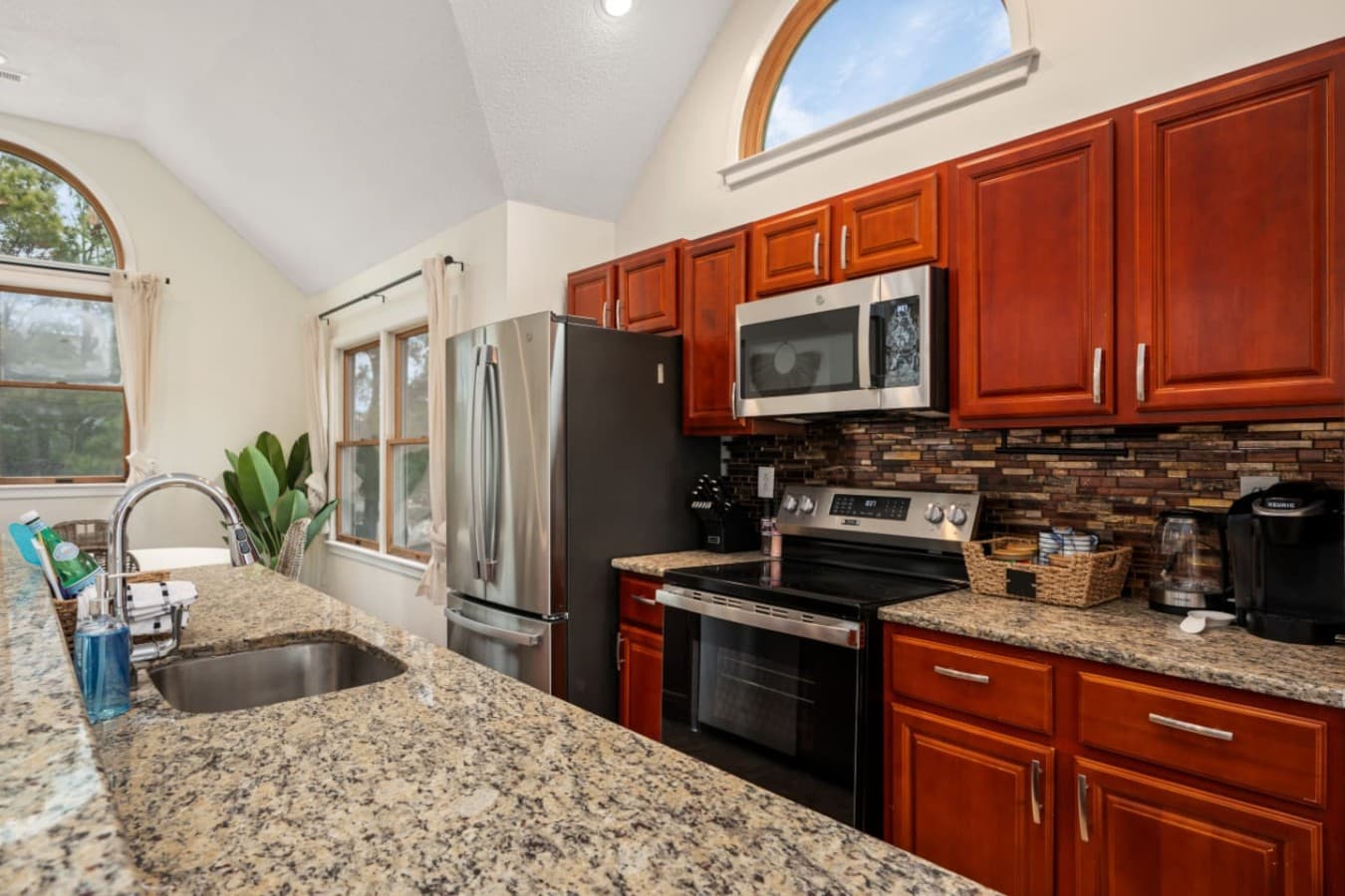 Kitchen w/ granite counters, cherry cabinetry, stainless appliances & arched windows above.