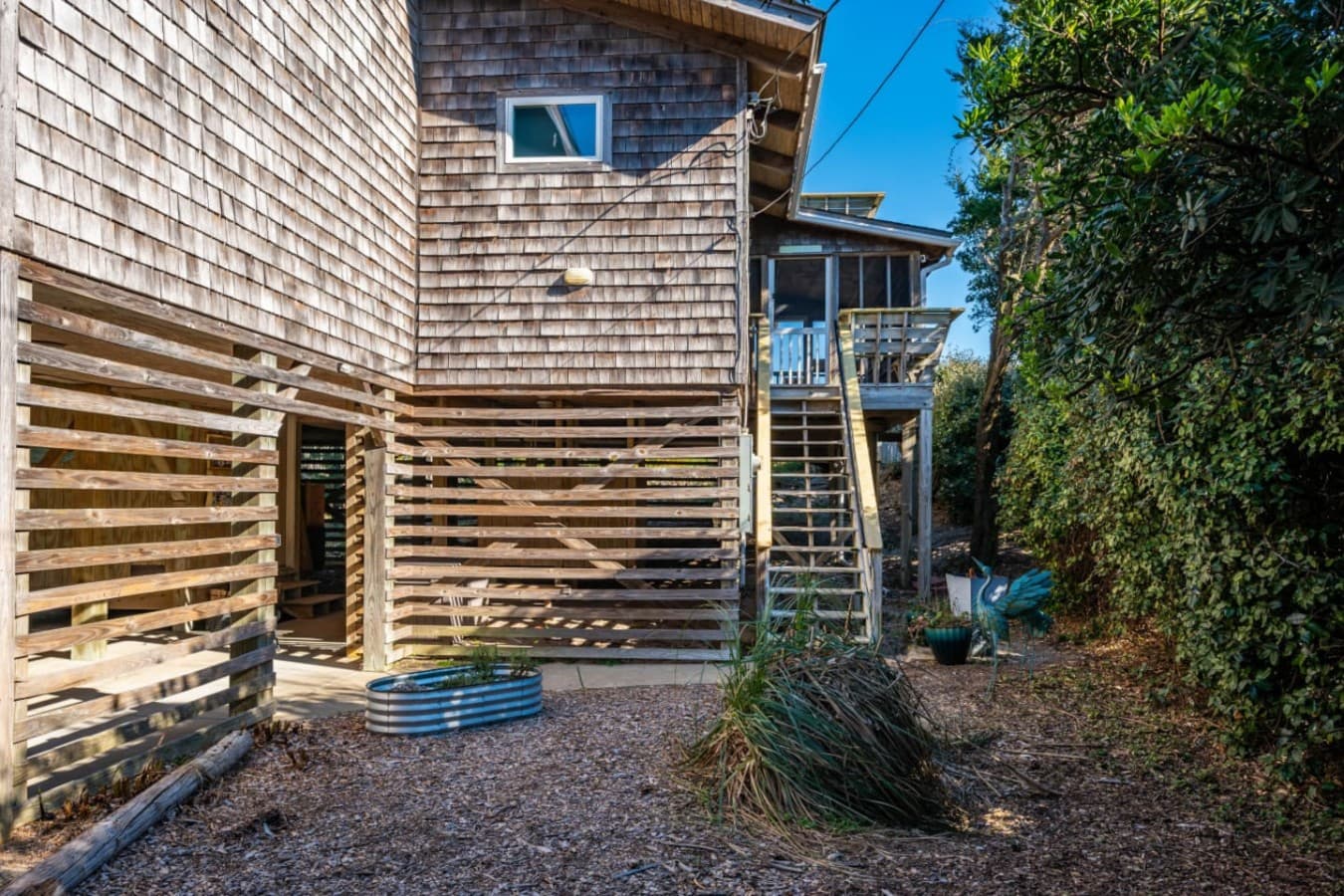 Sunlit exterior w/ elevated deck, wood siding, & shaded ground-level space framed by natural greenery.
